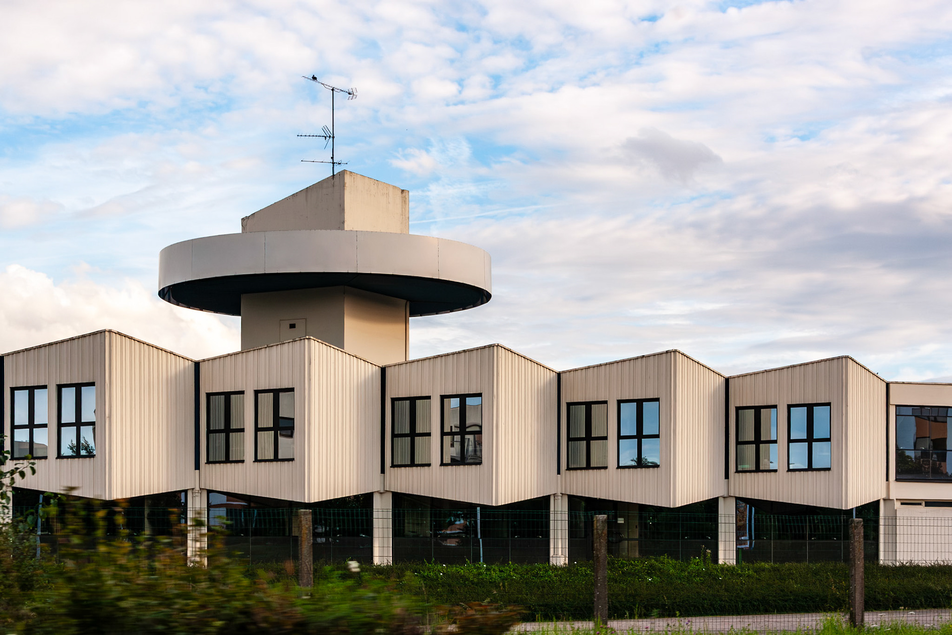 A contemporary commercial building in France featuring a distinctive jagged facade, large windows, and a central tower with circular signage under a blue sky with scattered clouds.