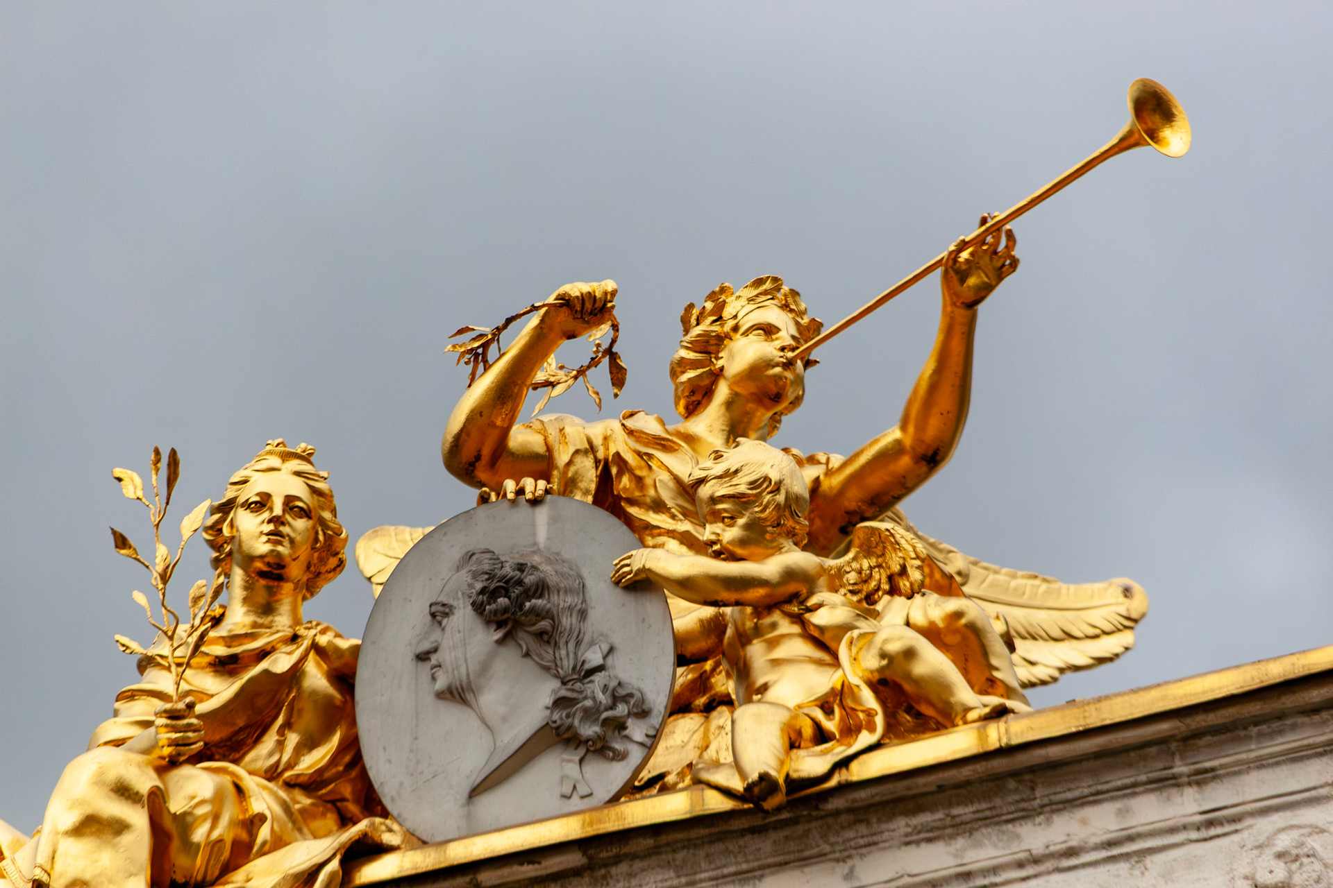 A detailed close-up of a gold-leafed sculptural arrangement at Place Stanislas in Nancy, France, featuring a winged figure blowing a trumpet, a seated woman holding an olive branch, and a cherub flanking a central stone medallion.