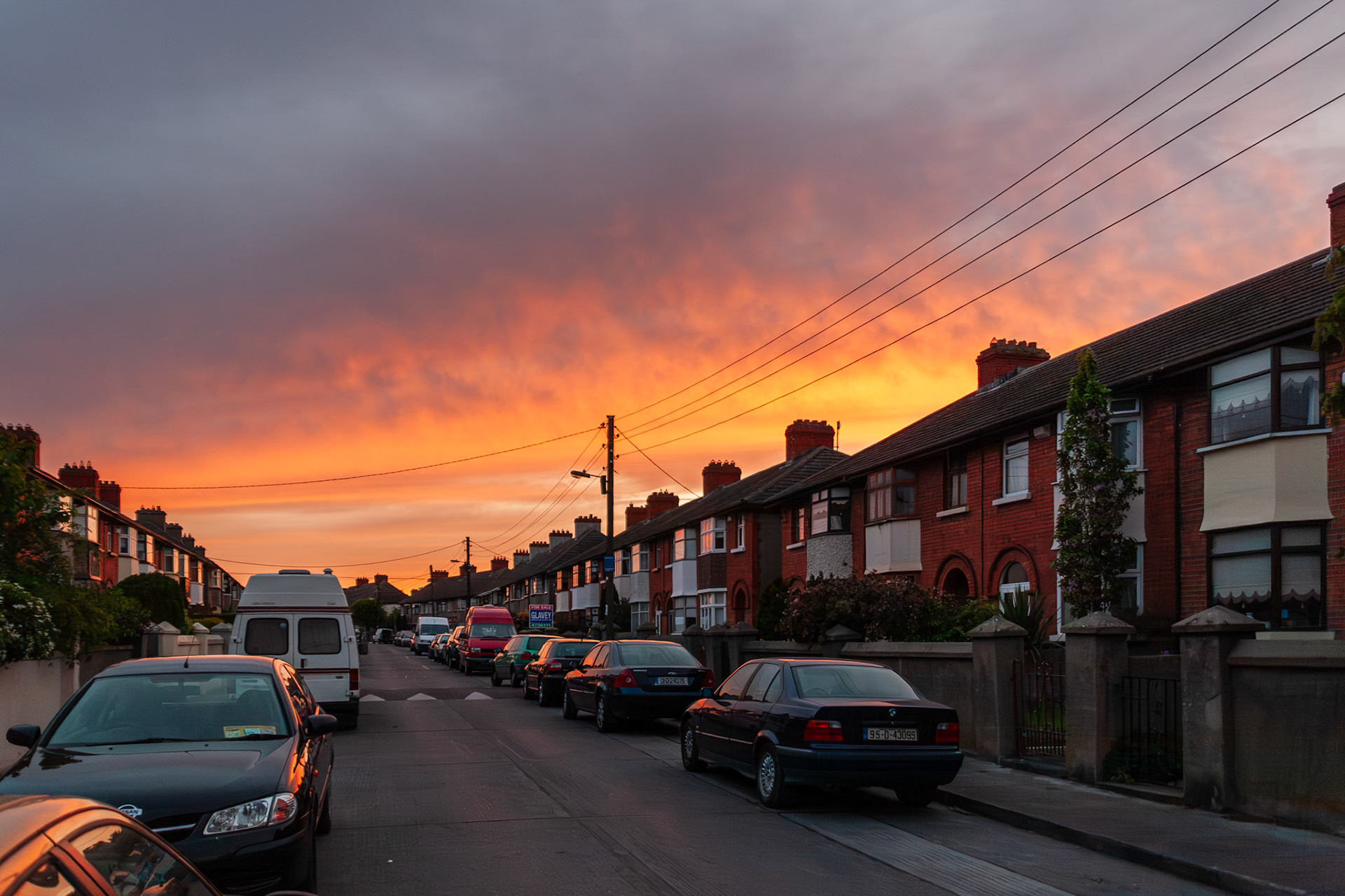 A vibrant orange and purple sunset fills the sky above a traditional terrace of red-brick houses and parked cars on a quiet residential street in Dublin, Ireland.