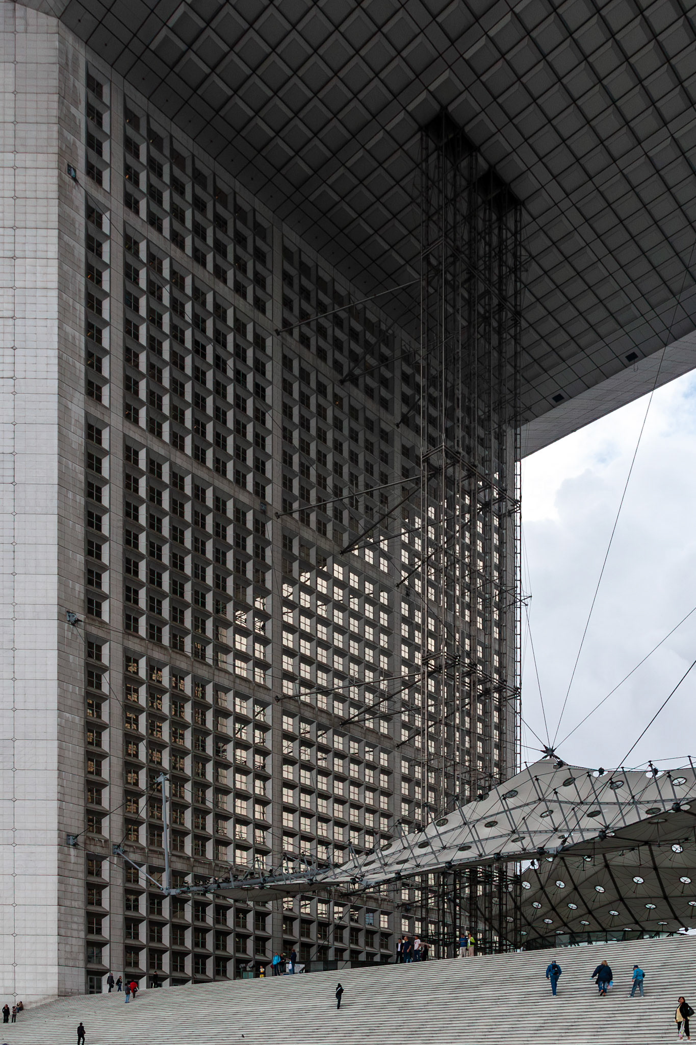 A striking low-angle view of a monumental modern building featuring a repetitive grid facade and a vast overhanging roof. The structure is complemented by a unique white geometric canopy and wide public stairs where people are seen walking, emphasizing the scale and contemporary design of the urban space.