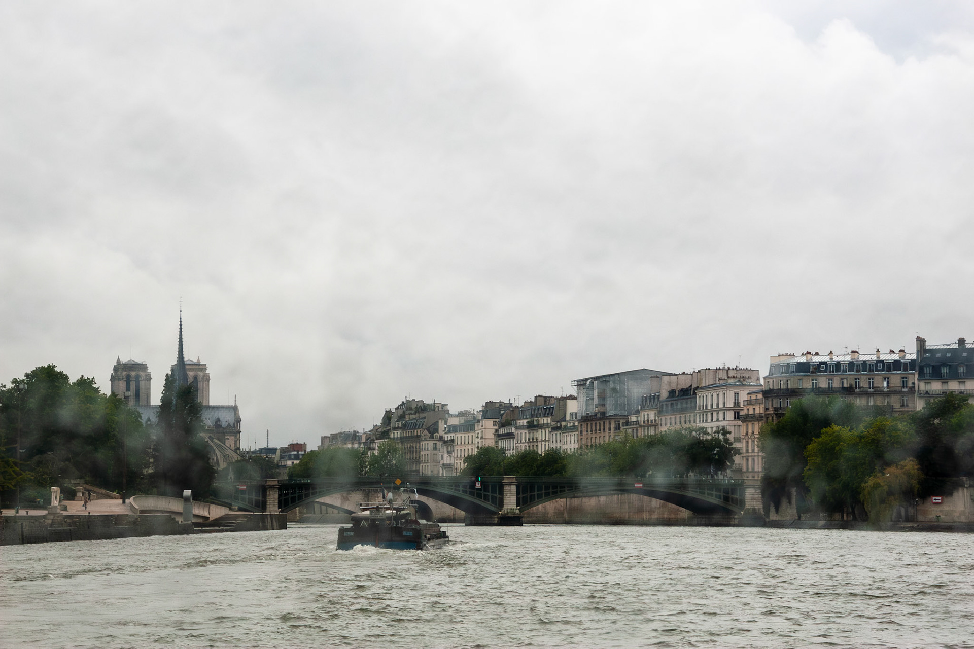 A wide-angle view from the water showing a boat navigating the Seine River in Paris, with a bridge in the mid-ground and the historic skyline, including the towers of Notre Dame Cathedral, under a heavy overcast sky.