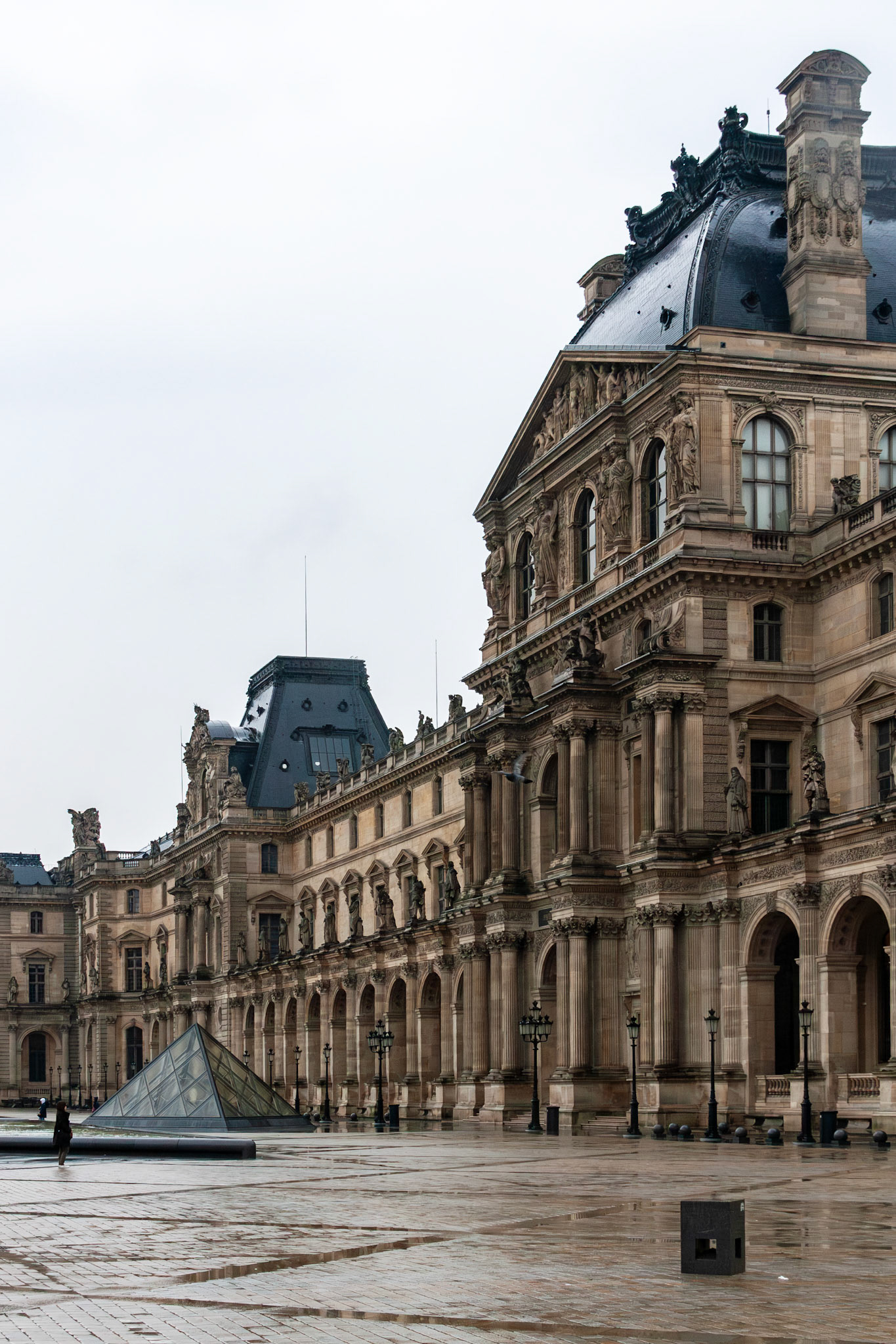 Wide view of the historic stone facade and courtyard of a world-renowned Parisian art museum, showcasing classical French Renaissance architecture and modern glass structures.