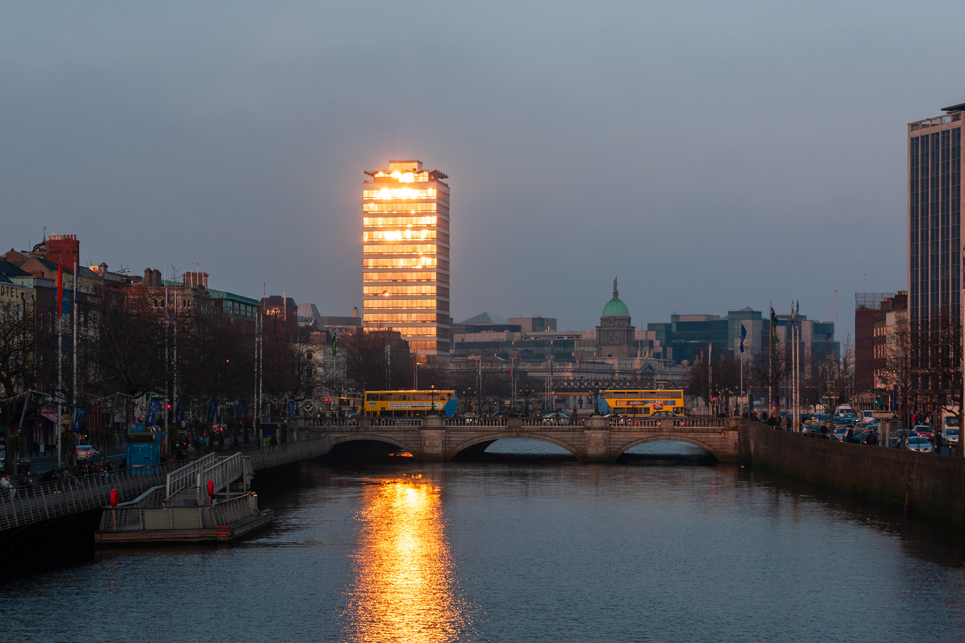 The setting sun reflects brilliantly off the glass facade of Liberty Hall skyscraper, overlooking O'Connell Bridge and the River Liffey in Dublin city center at dusk.