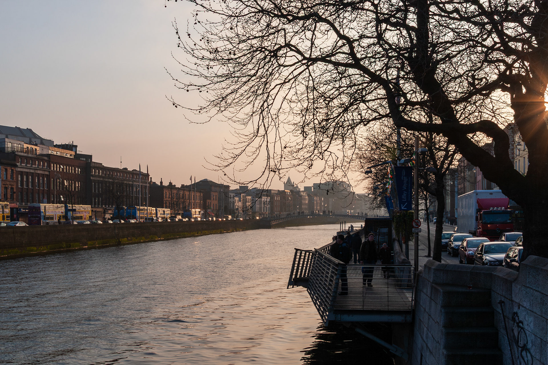 A scenic view of the River Liffey in Dublin, Ireland, during sunset, featuring the city quays, silhouetted winter trees, and a boardwalk with pedestrians under a warm evening sky.