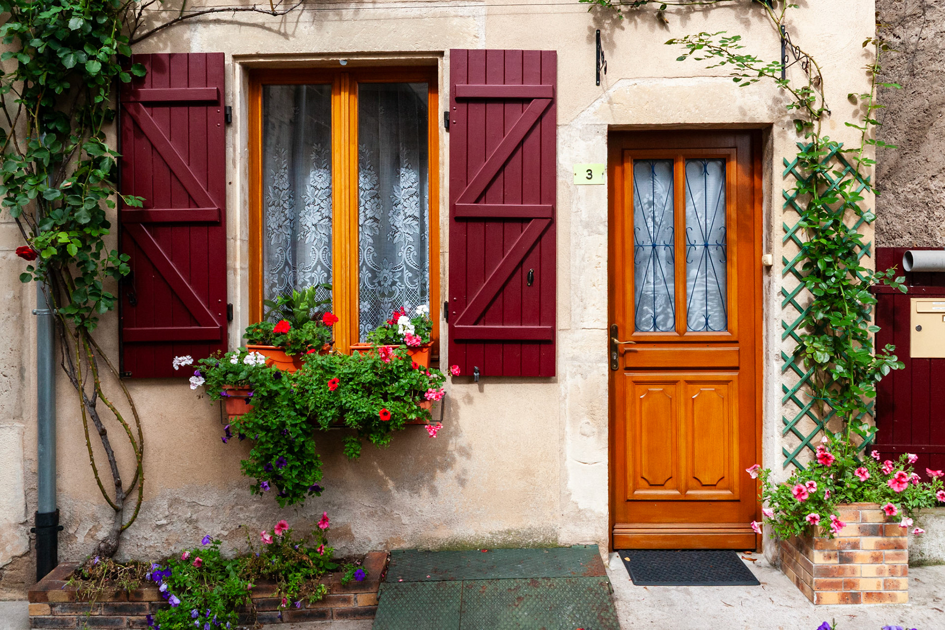 A charming and picturesque entrance to a traditional French home, featuring a wooden door, bright red window shutters, lacy curtains, and vibrant flower boxes with climbing green vines on a stone facade.