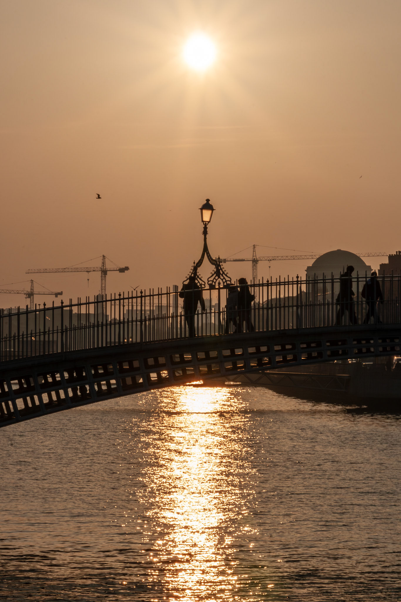 A golden sunset over the River Liffey in Dublin, Ireland, featuring the iconic Ha'penny Bridge silhouetted against a bright sun with pedestrians crossing and sunlight reflecting on the water.