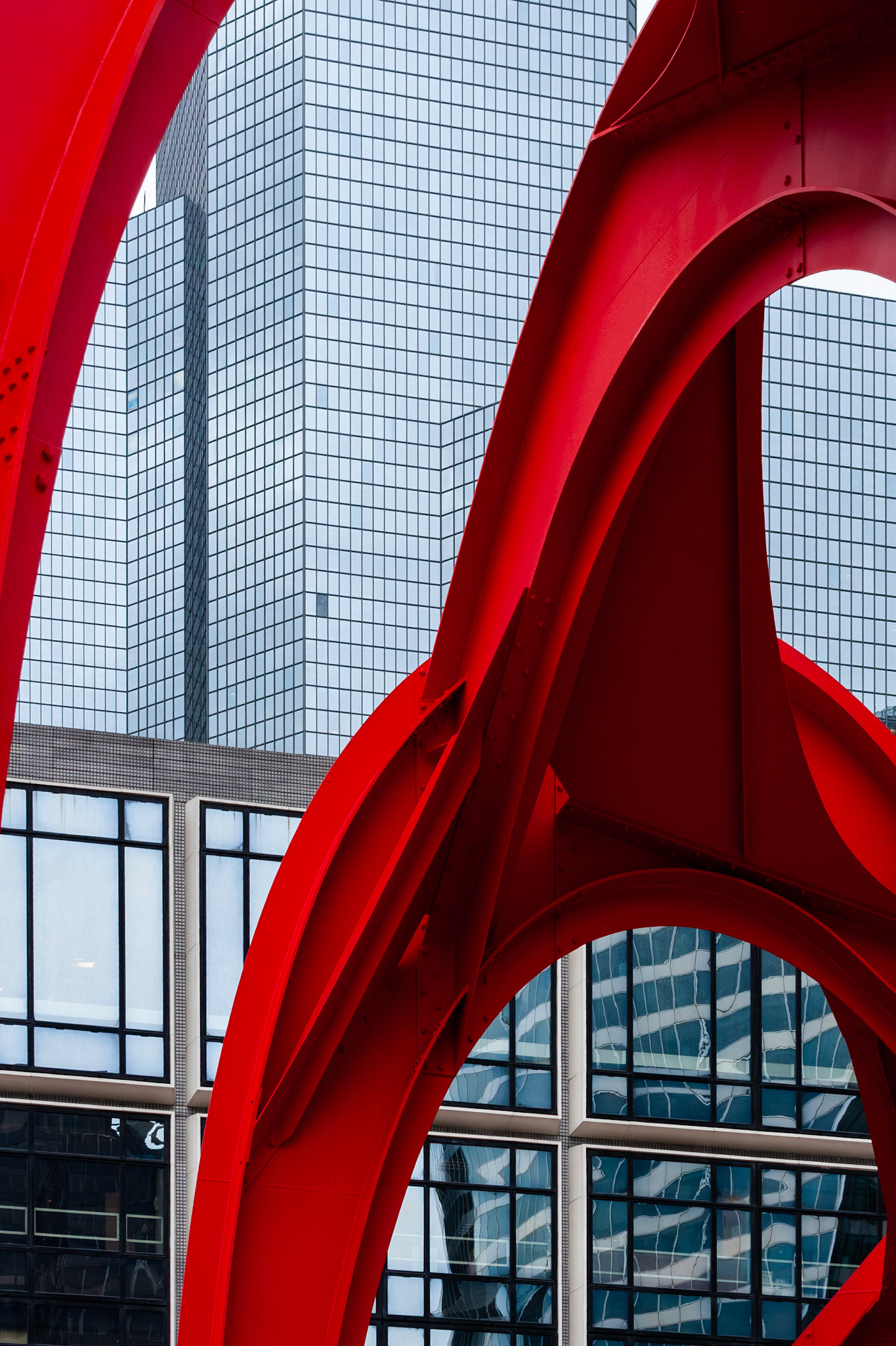 A bold red metal sculpture with curved, arching lines frames a background of modern glass skyscrapers. The contrast between the bright primary color of the industrial art and the repetitive blue-gray grid of the office building windows creates a dynamic architectural composition.