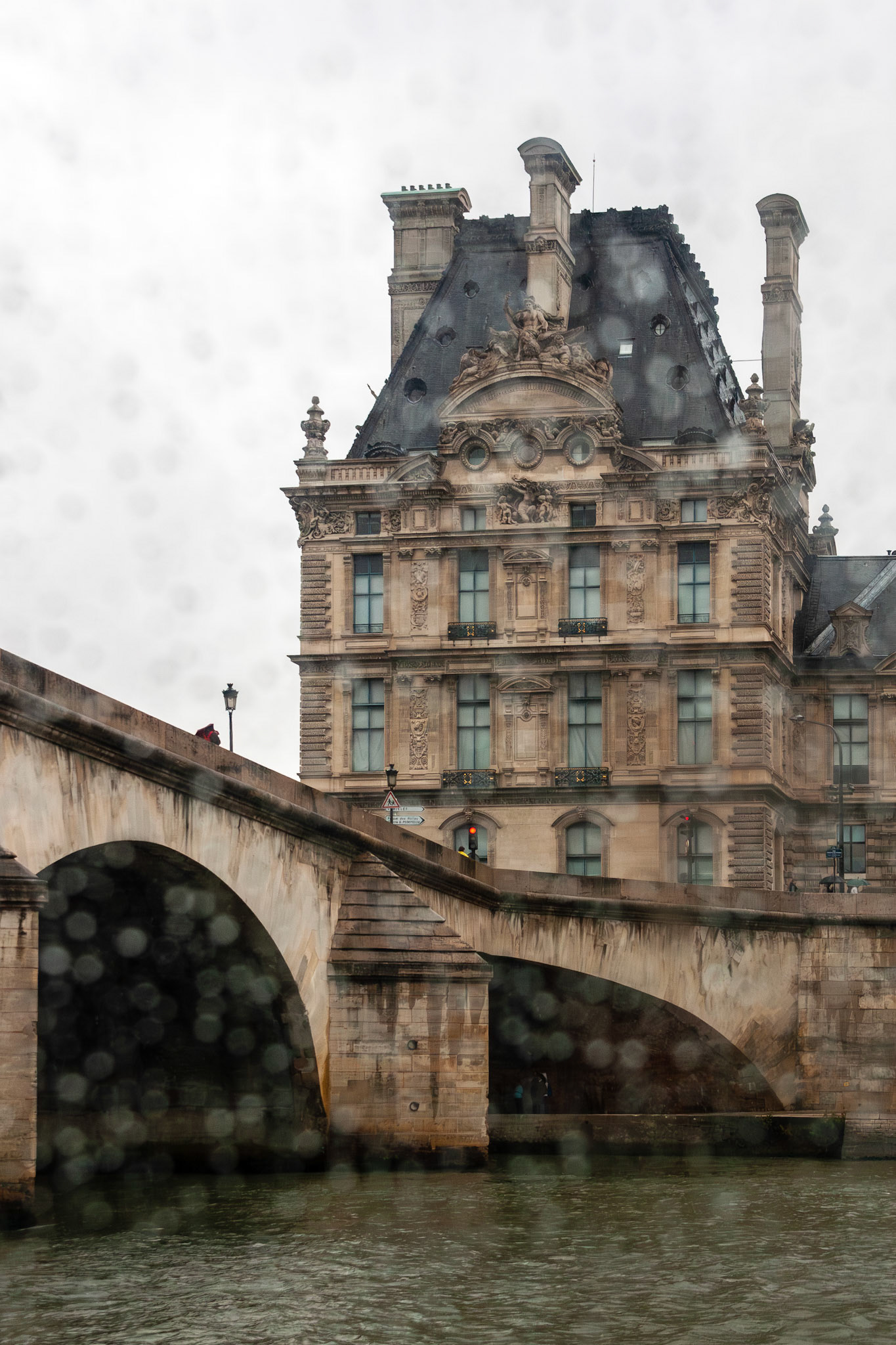 A view of a classic French limestone building and stone arch bridge along the Seine River in Paris, seen through a window with soft raindrops and bokeh blurring the foreground for a moody atmosphere.