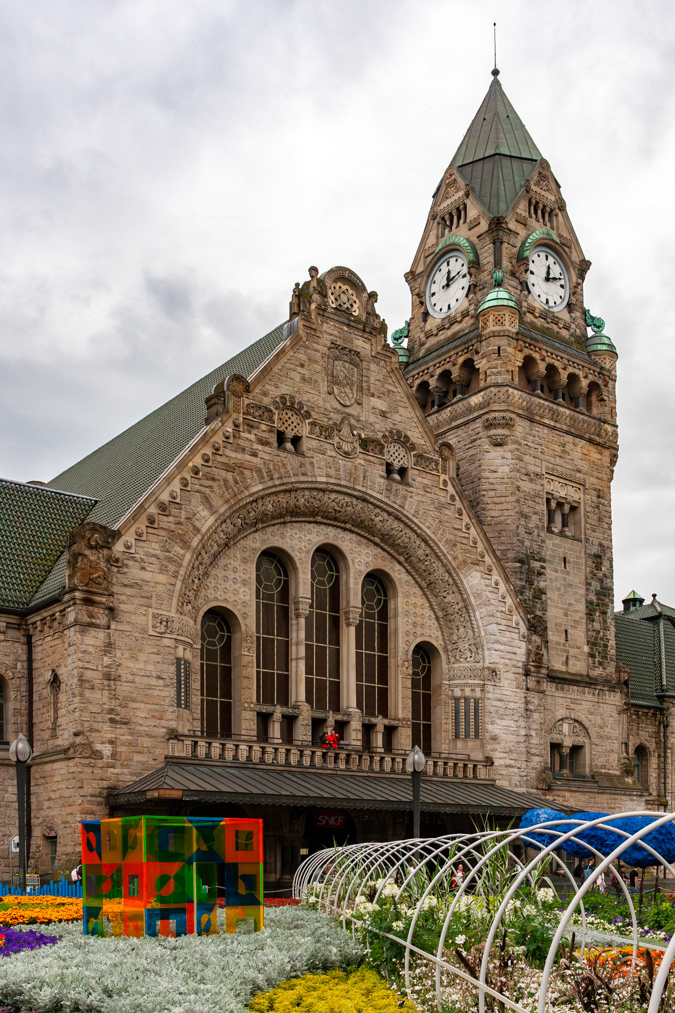 The Neo-Romanesque facade and ornate clock tower of the historic Gare de Metz-Ville railway station, viewed from a landscaped plaza featuring colorful flower beds and modern art installations in Metz, France.