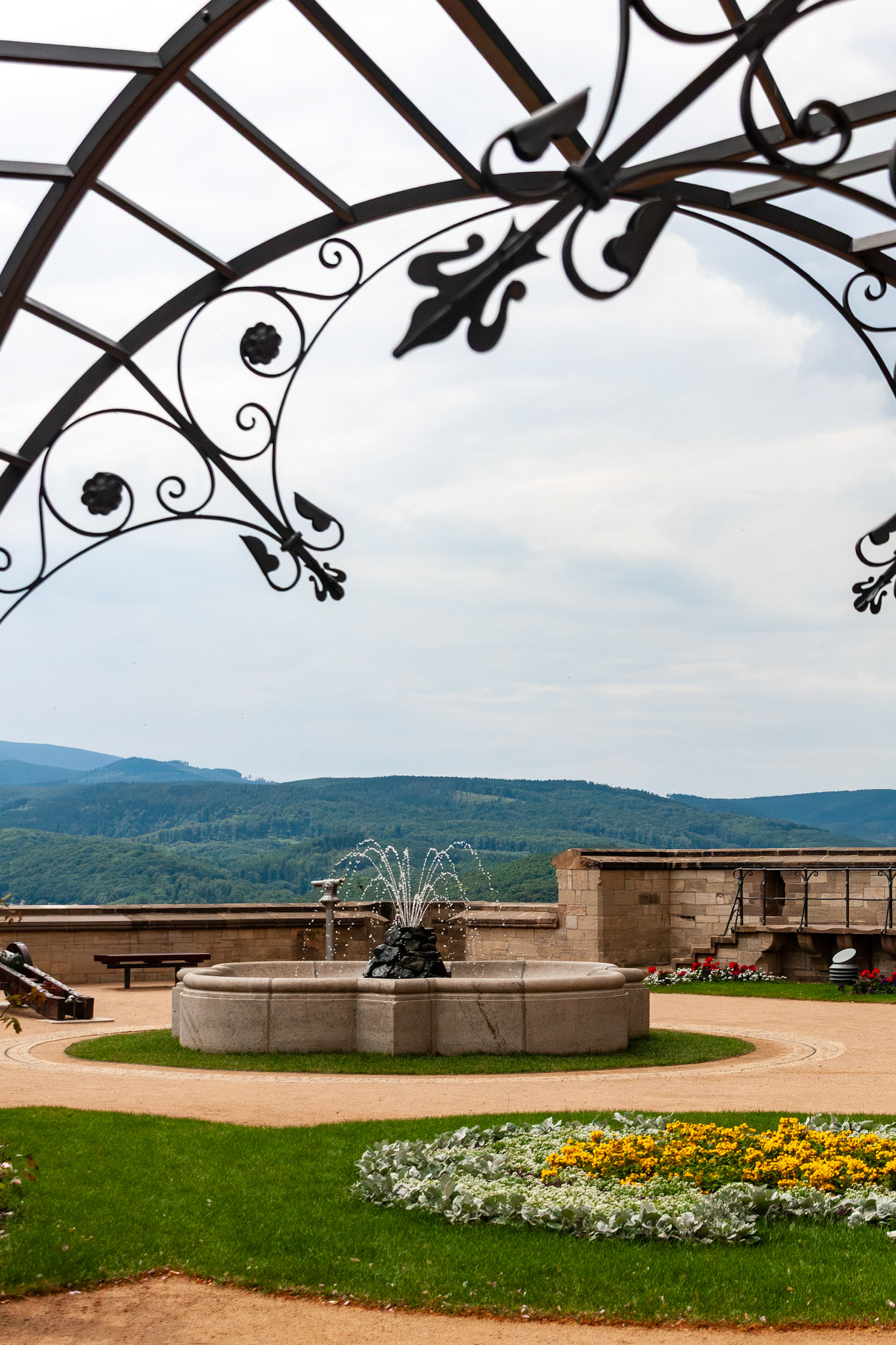 An elegant stone fountain sits in a manicured terrace garden at Wernigerode Castle, framed by a decorative wrought-iron arbor with a sweeping view of the Harz mountains in Germany.