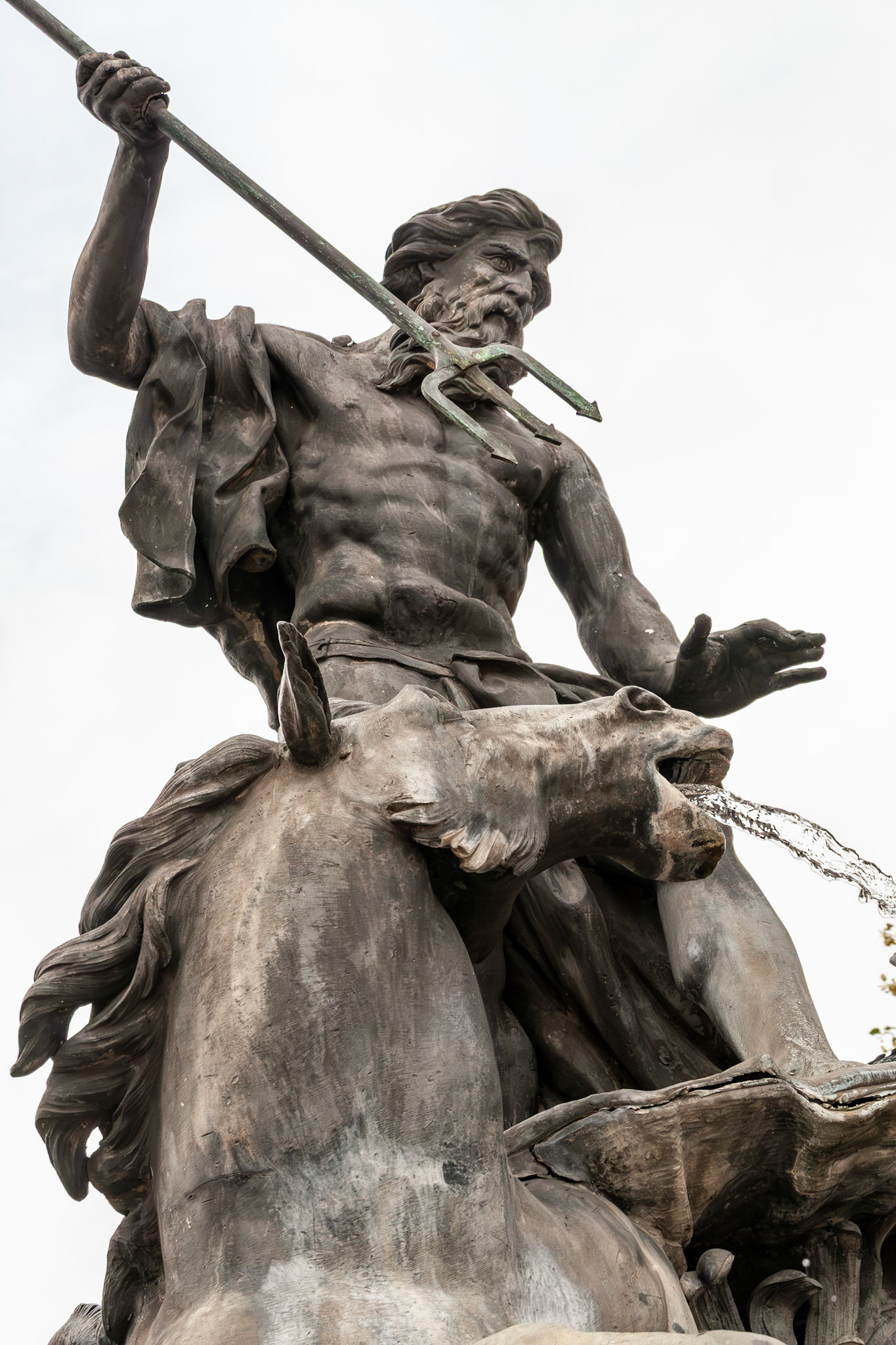 A low-angle, detailed shot of the central bronze sculpture from the Fountain of Neptune in Nancy, France, depicting a muscular Neptune wielding a trident while taming a water-spouting sea horse.