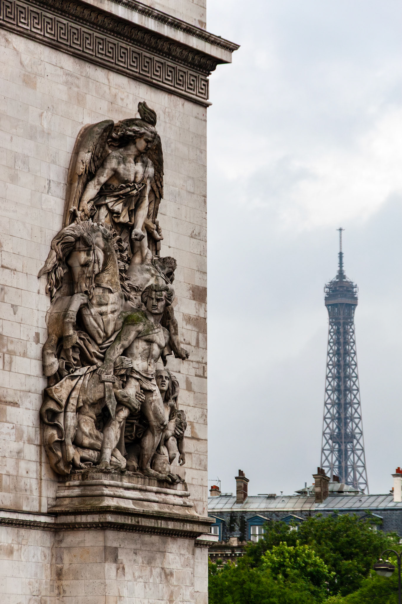 A vertical composition featuring a detailed limestone relief sculpture on the facade of the Arc de Triomphe, with the iconic silhouette of the Eiffel Tower visible in the distance under a soft, overcast Parisian sky. The image highlights the juxtaposition of two of France's most famous architectural landmarks.