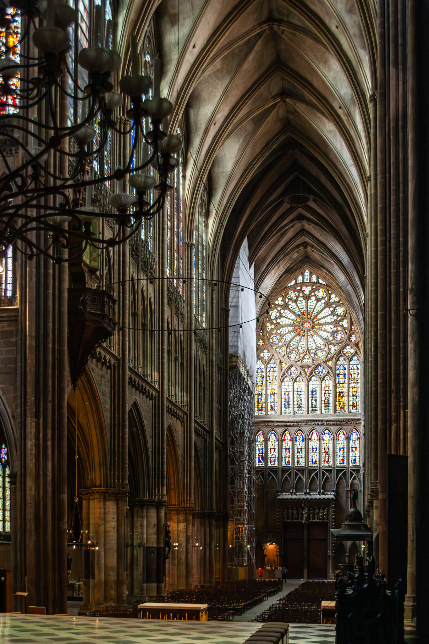 An interior view of the Saint Stephen Cathedral (Cathédrale Saint Étienne) in Metz, France, showcasing the soaring Gothic ribbed vaults, massive stone columns, and the magnificent stained glass rose window above the transept.