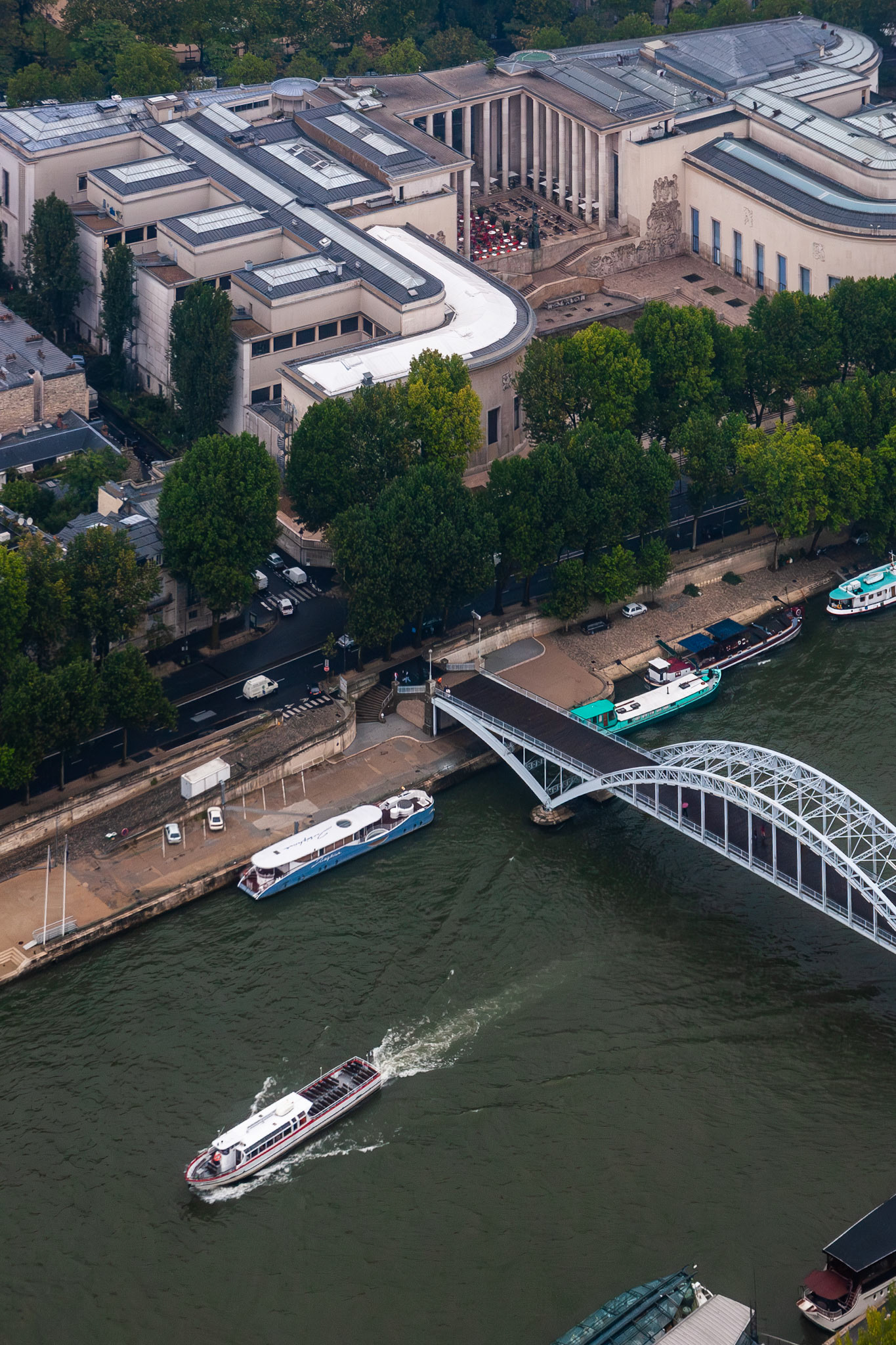 A high-angle perspective of the Palais de Tokyo museum on the banks of the Seine River in Paris, featuring a modern pedestrian bridge and tourist boats navigating the water.