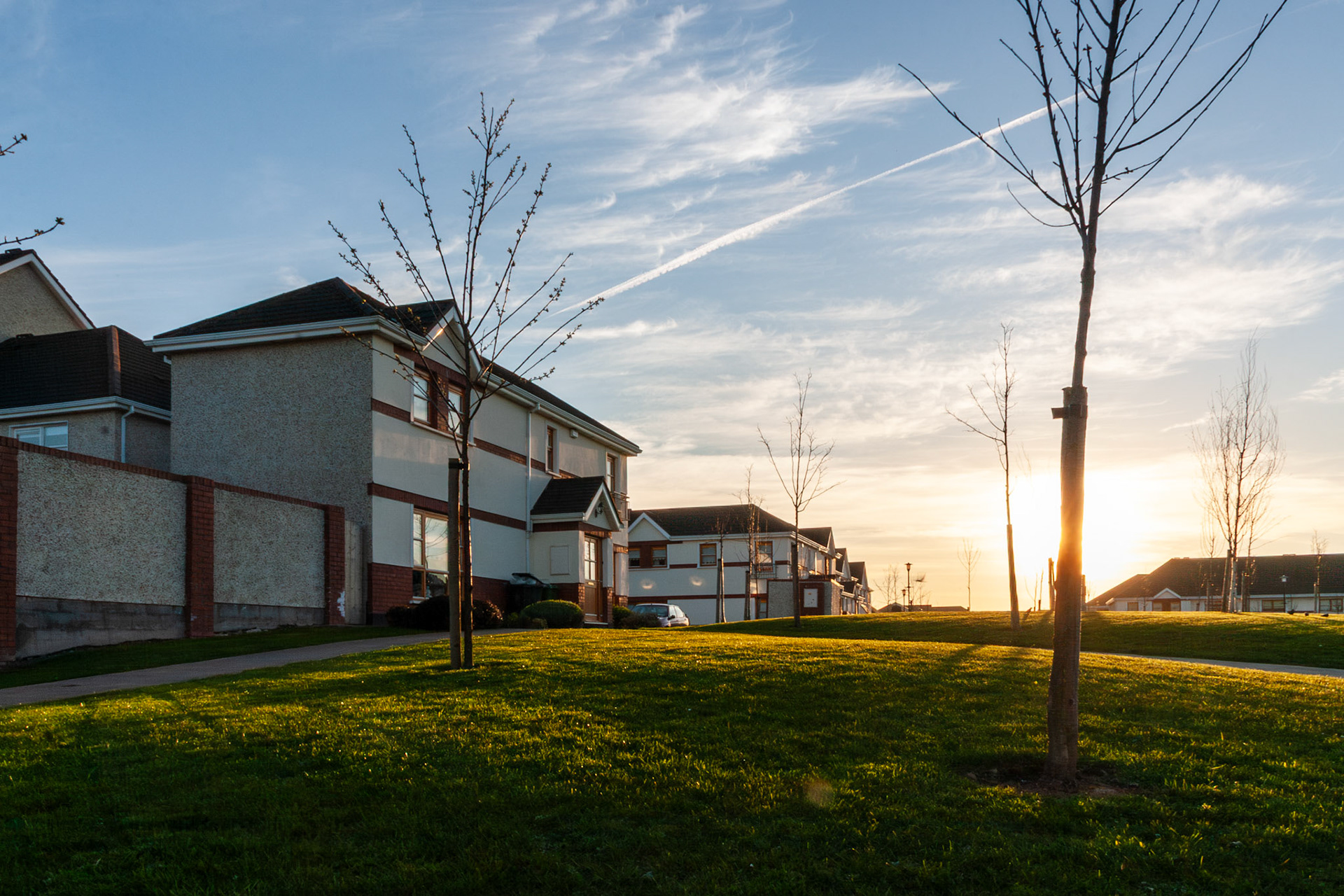 A peaceful sunset view of a contemporary housing estate in Dublin, Ireland, featuring modern detached homes, young trees, and a vibrant green lawn illuminated by warm evening sunlight under a blue sky with wispy clouds.