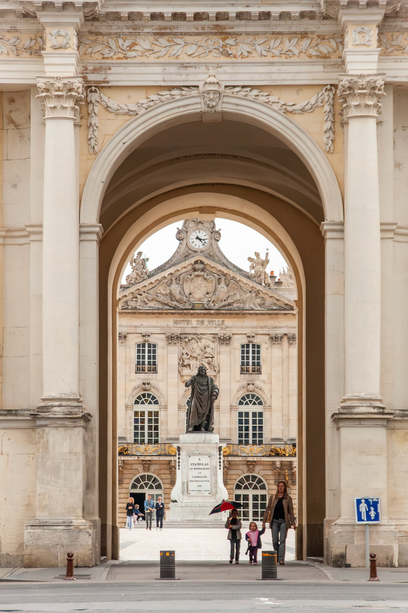 A vertical perspective through the grand neoclassical archway of the Arc Héré, framing the bronze statue of Stanislas Leszczynski and the ornate facade of the Hôtel de Ville (City Hall) in Nancy, France.