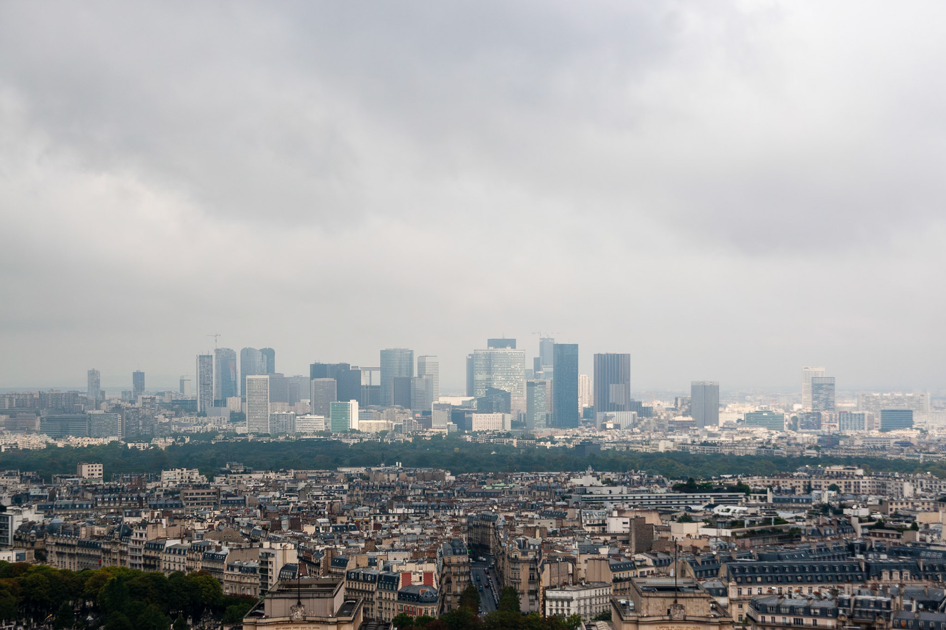 A panoramic view of the modern skyscrapers in the La Defense business district under a moody, overcast sky. The foreground features the classic Haussmann-style architecture of Paris, highlighting the contrast between the historic city and the contemporary financial hub in the distance.