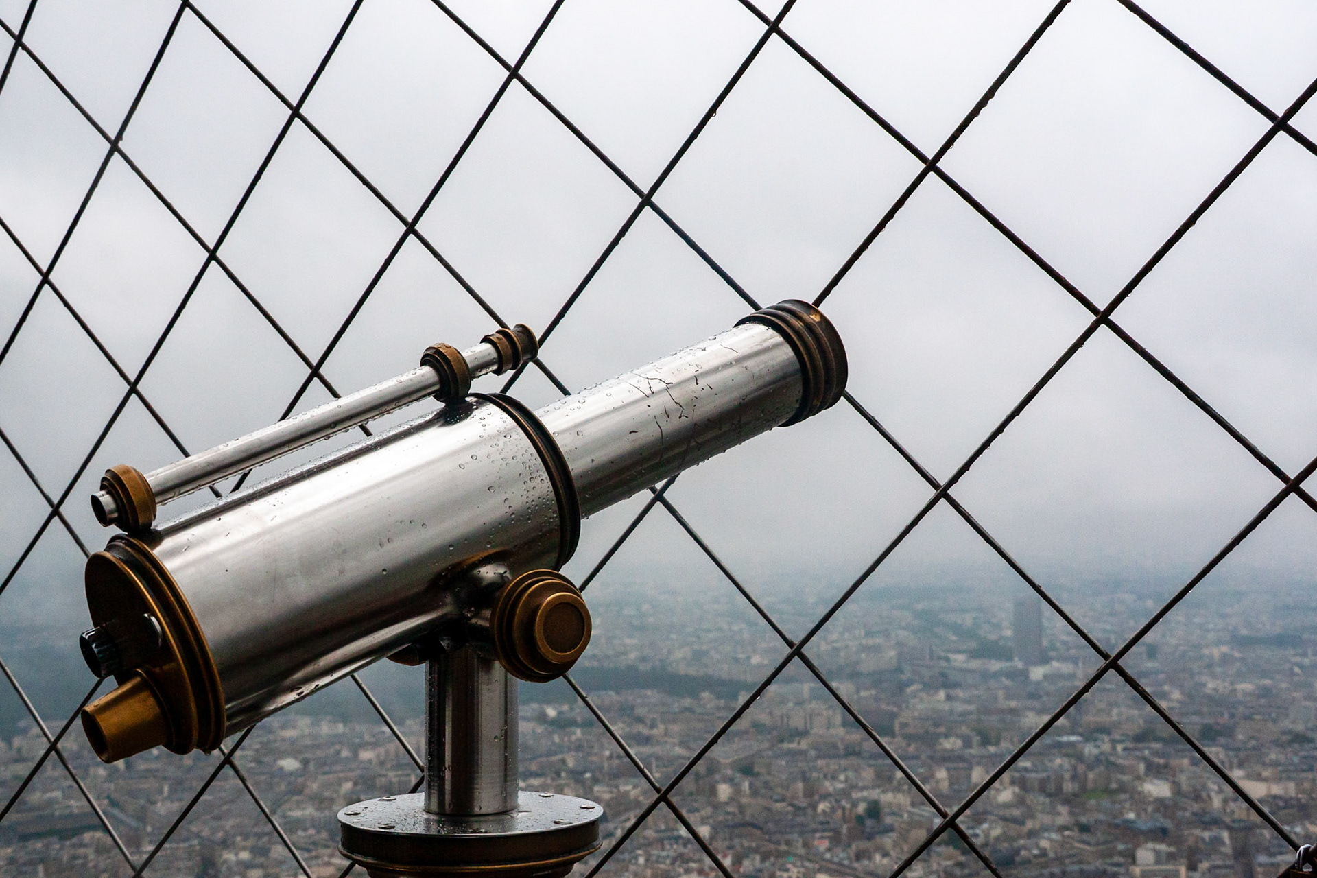 A close-up side view of a classic silver and brass coin-operated telescope mounted on the Eiffel Tower, covered in raindrops against a foggy Paris city backdrop seen through a safety wire mesh.