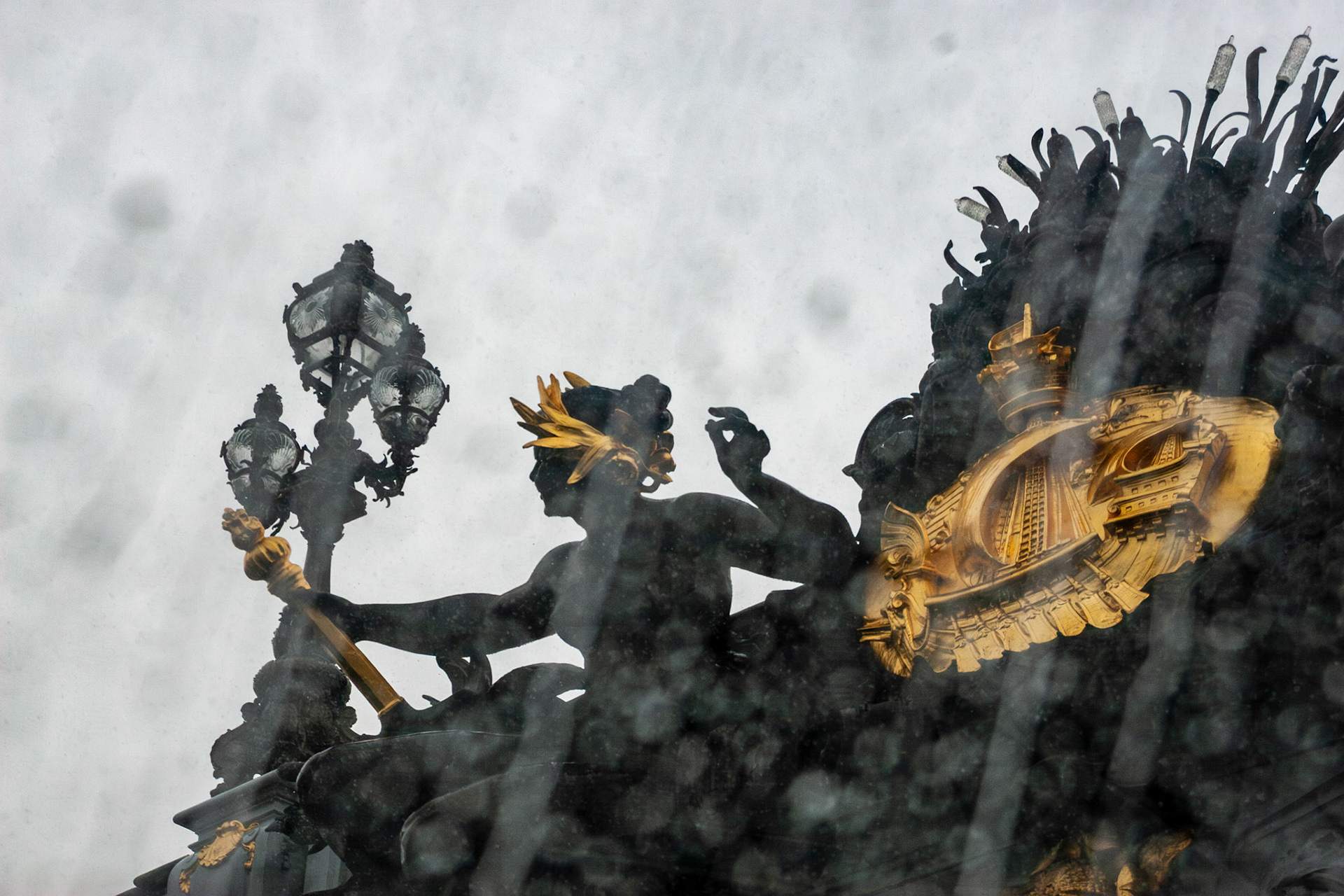 A close-up view of the intricate bronze and gold-leaf sculptures on the Pont Alexandre III bridge in Paris, France, captured through a rain-streaked window or lens, creating a moody, atmospheric urban scene.