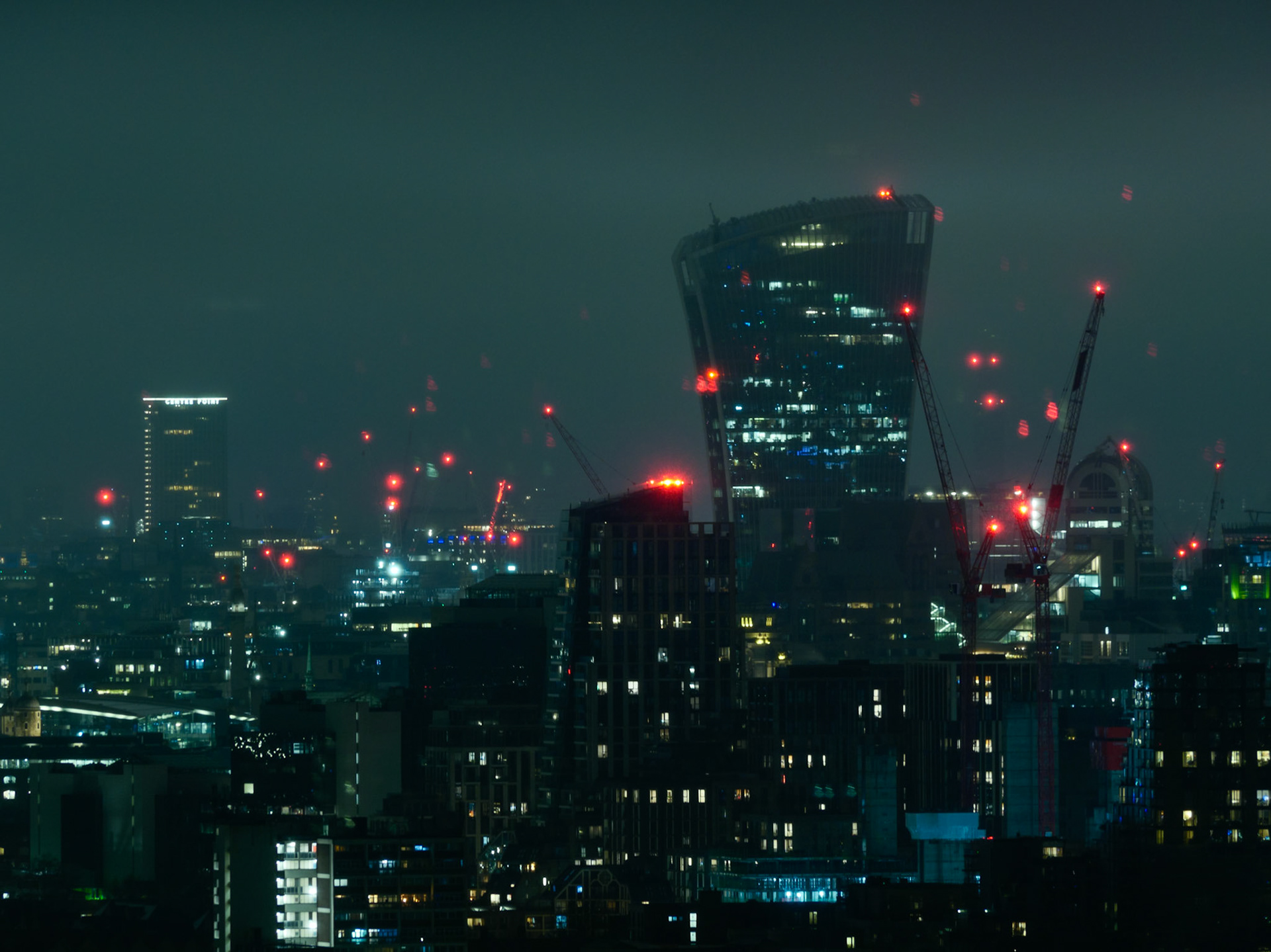 A moody, futuristic cityscape of London at night, featuring the illuminated Walkie Talkie building and construction cranes against a dark, misty sky filled with red aviation warning lights.