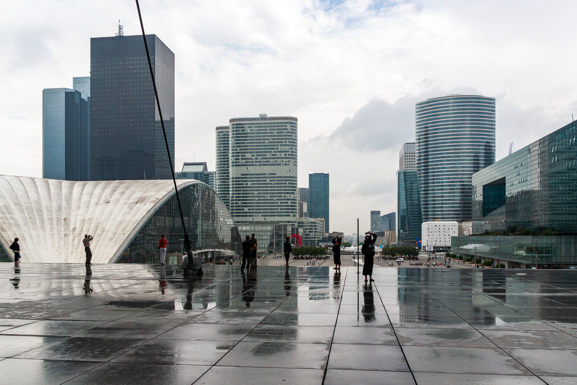 A wide-angle view of a contemporary business district featuring various glass skyscrapers and a curved architectural structure. Pedestrians are scattered across a large, wet stone plaza that creates dark, mirror-like reflections under an overcast sky, highlighting the symmetry and urban atmosphere.