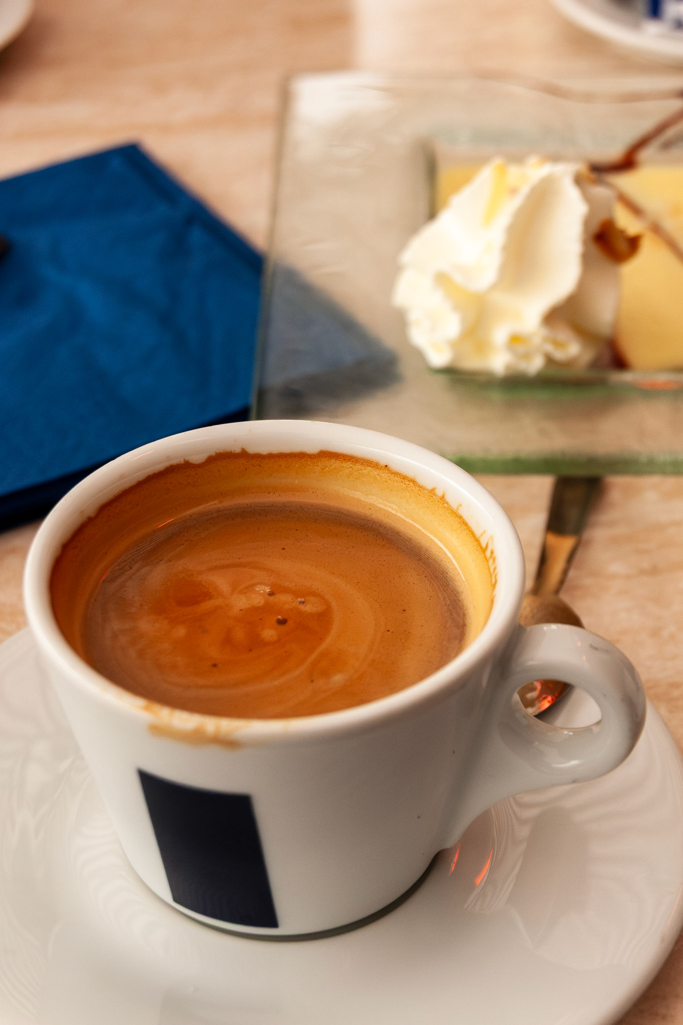 A detailed close-up shot of a white ceramic cup of espresso with rich crema, accompanied by a gourmet dessert topped with whipped cream on a marble tabletop at an outdoor café in Nancy, France.