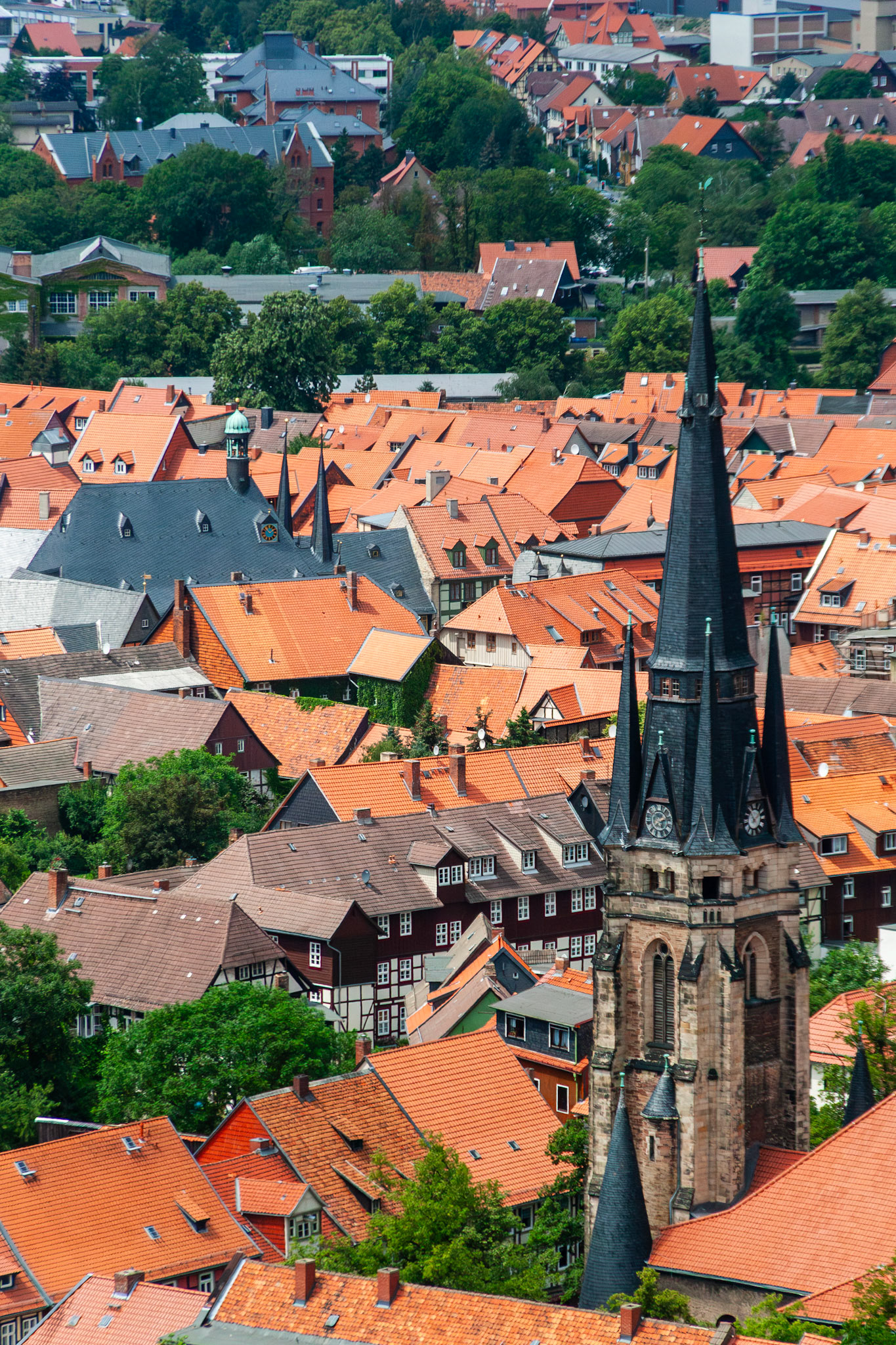 A high-angle view of Wernigerode, Germany, featuring a prominent dark Gothic church spire with a clock tower set against a sea of traditional orange-tiled rooftops and timber-framed buildings.