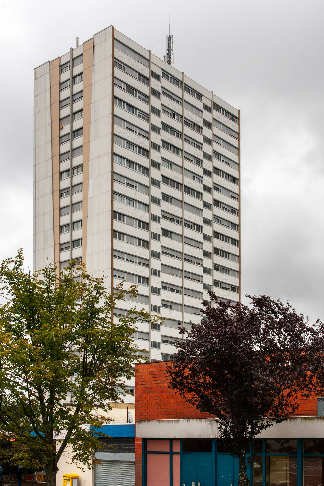 A tall, multi-story residential apartment block with a white and tan facade stands under an overcast sky in Nancy, France, with urban greenery and the "Salle Louis" building in the foreground.