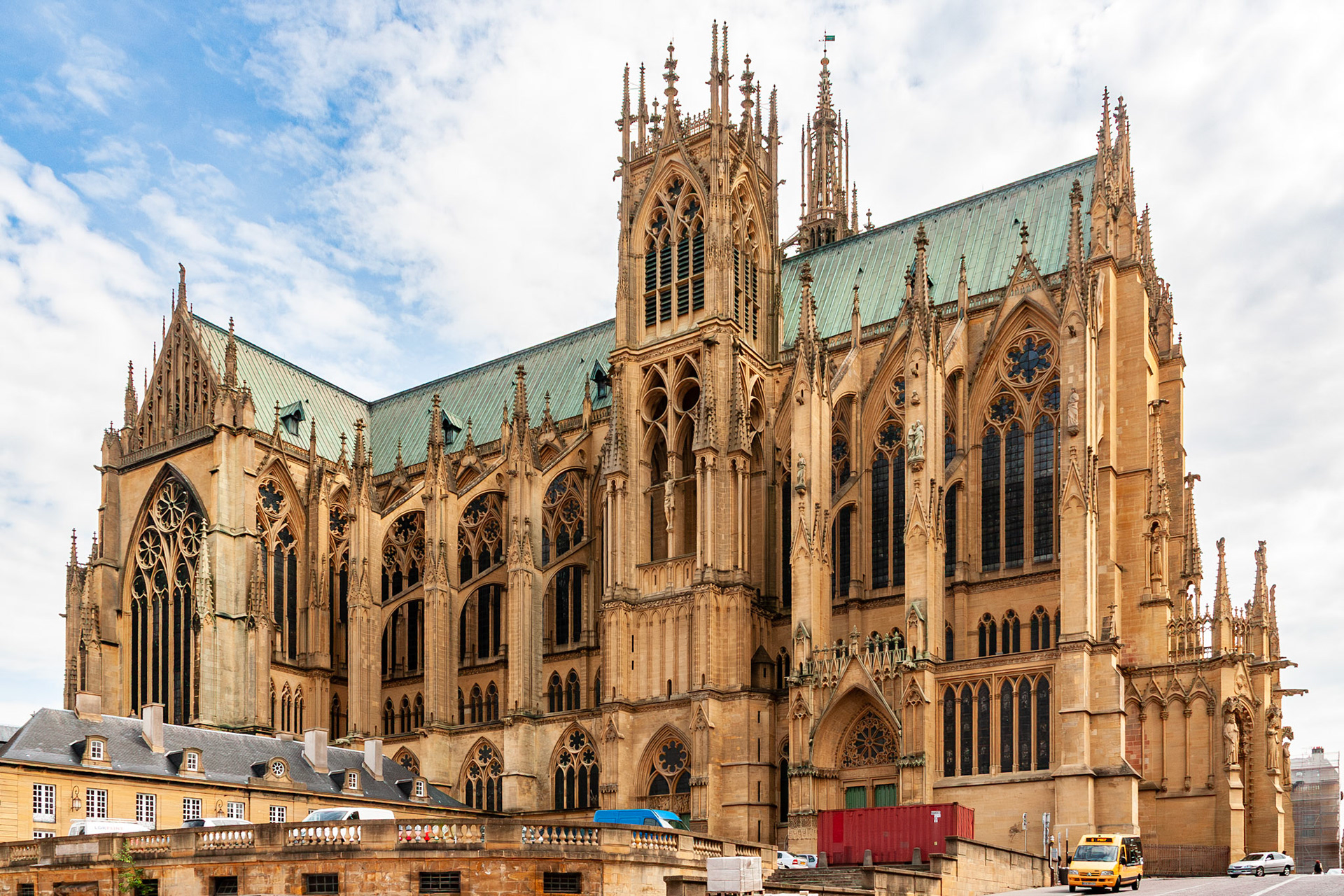 A full architectural exterior view of the Cathédrale Saint-Étienne de Metz, showcasing its golden Jaumont limestone, intricate Gothic flying buttresses, and expansive green-copper roof under a bright sky in the Grand Est region.