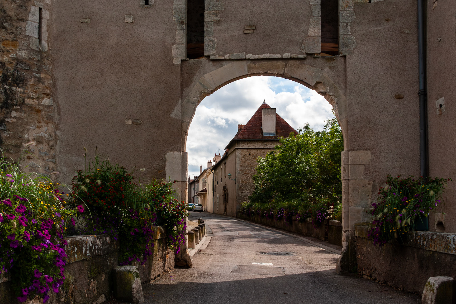 A view through an ancient stone archway leading into a quiet village street. The scene features weathered masonry walls, vibrant purple flowers in the foreground, and traditional French houses under a cloudy sky.