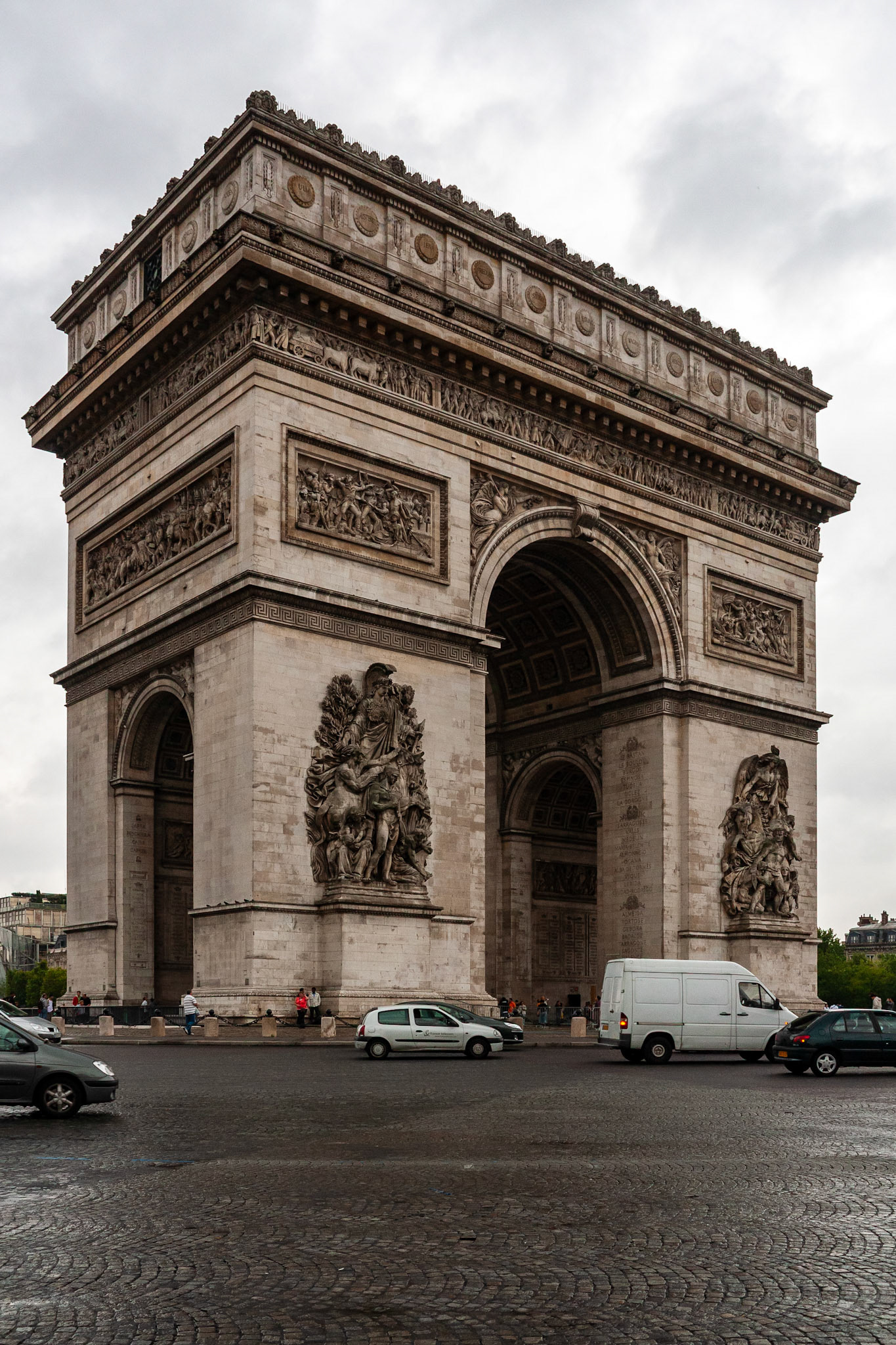 A wide shot of the iconic Arc de Triomphe de l'Étoile in Paris under an overcast sky. The image captures the neoclassical triumphal arch and its intricate relief sculptures, with city traffic and cars passing through the Place Charles de Gaulle in the foreground.