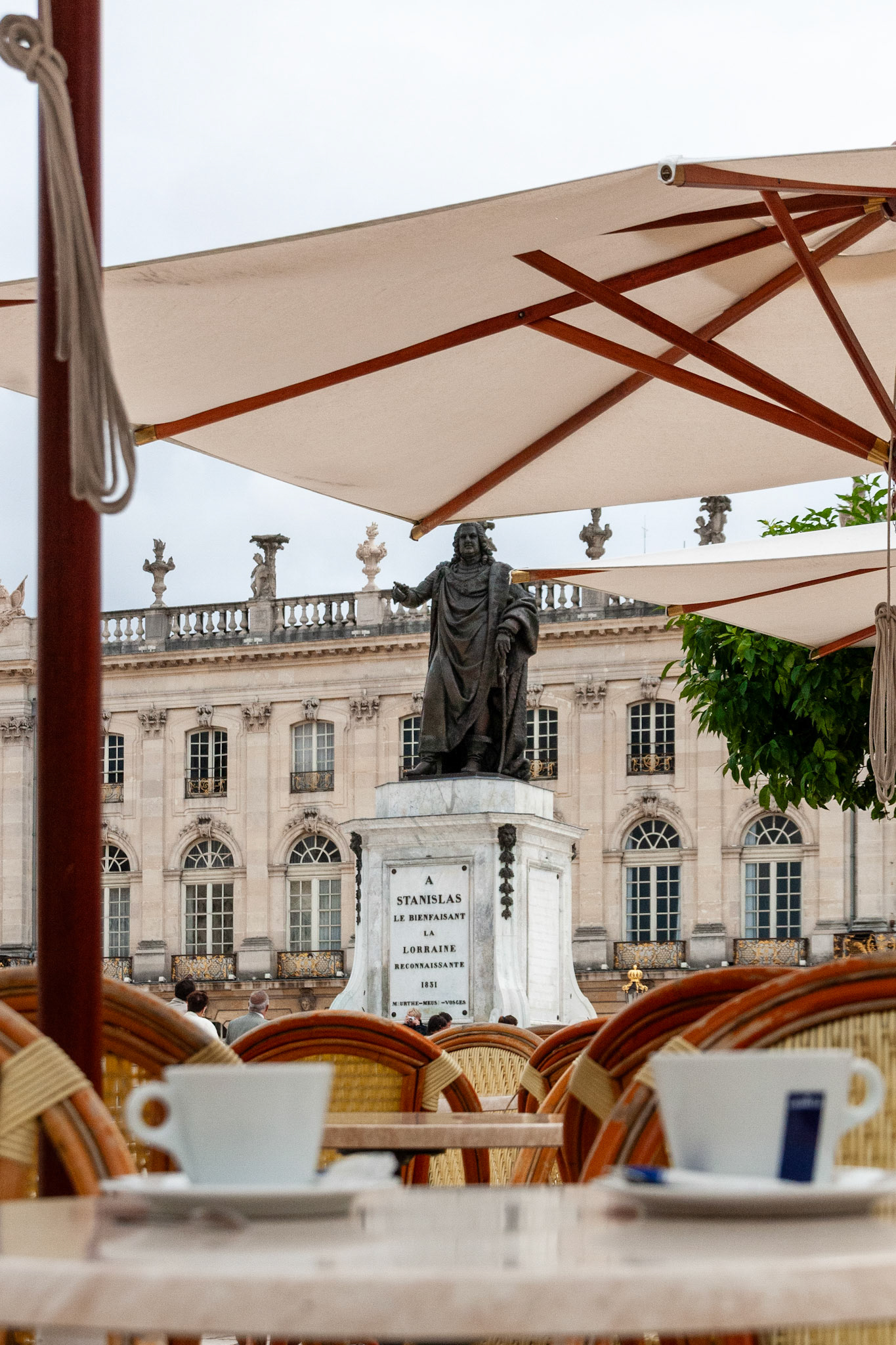 A relaxed view from a café terrace at Place Stanislas in Nancy, France, featuring white coffee cups and wicker chairs in the foreground, with the historic bronze statue of Stanislas I and grand neoclassical architecture in the background.