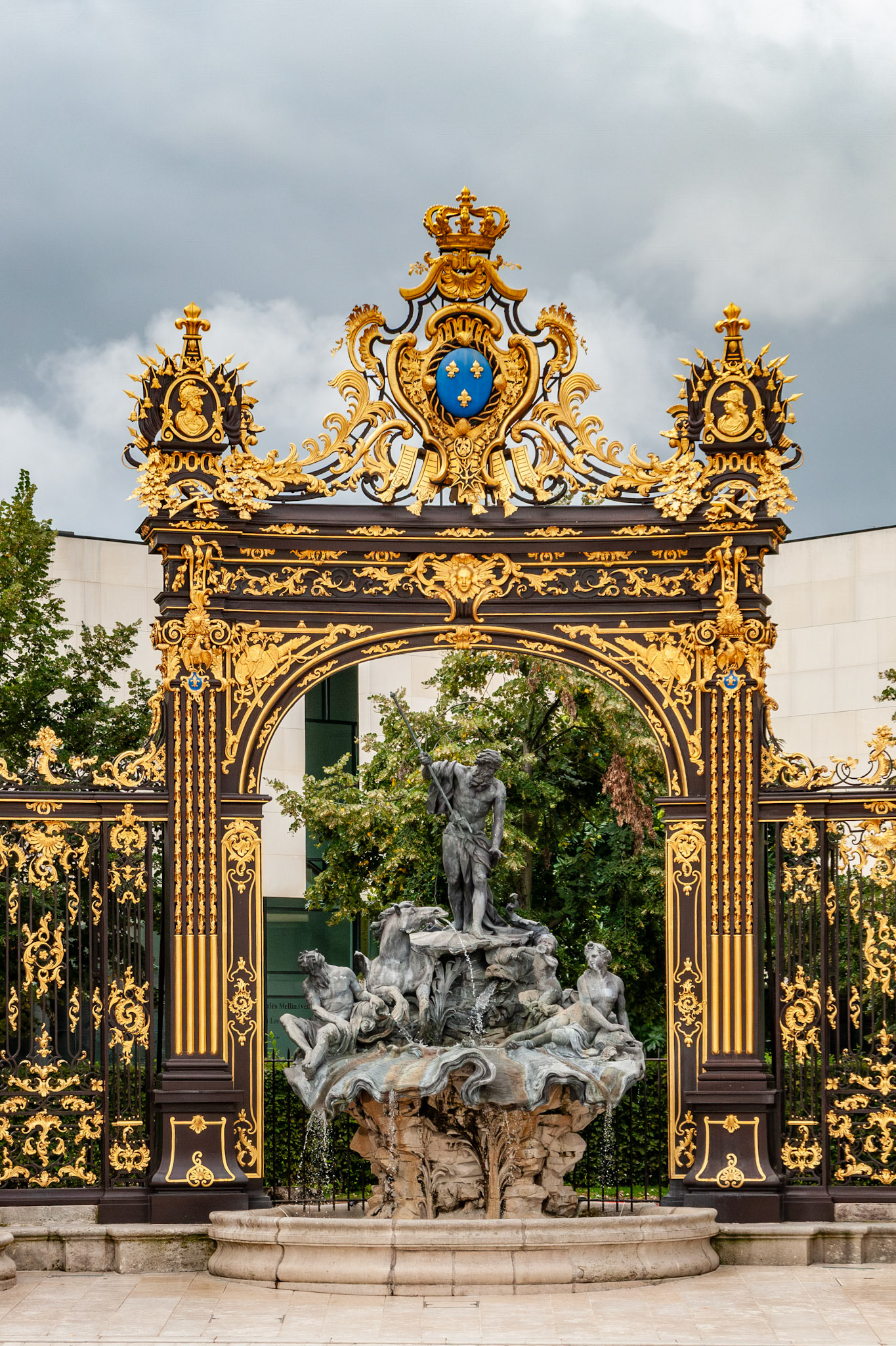 A majestic view of the Fountain of Neptune, featuring lead sculptures of Neptune with a trident, framed by the world-famous black and gold wrought-iron gates in the historic Place Stanislas of Nancy, France.