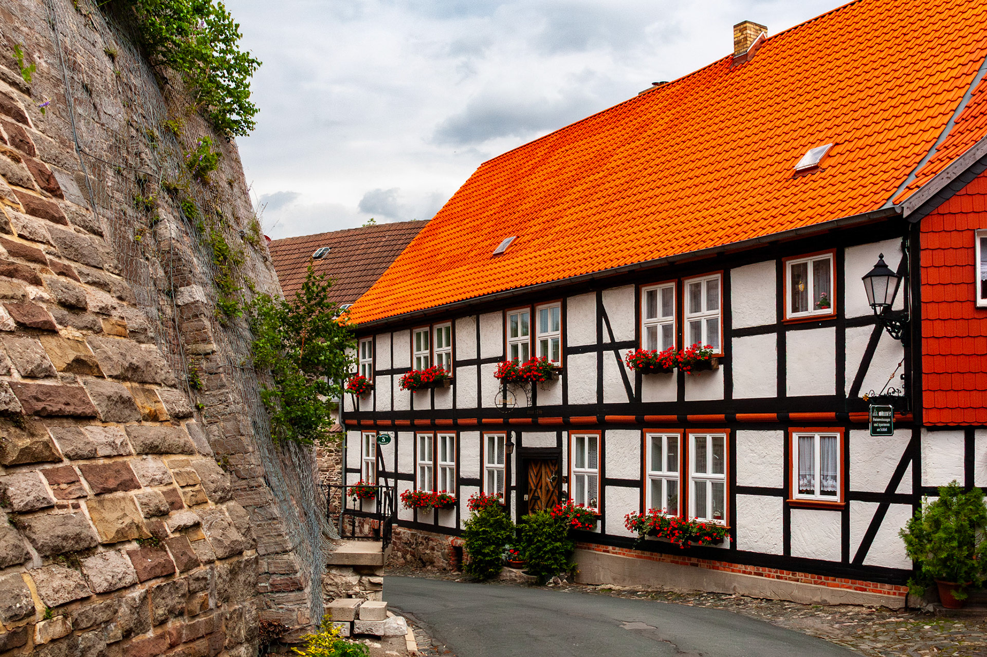A beautifully preserved half-timbered house with a bright orange tiled roof and red flower boxes sits along a narrow, winding street beneath a historic stone wall in Wernigerode, Germany.