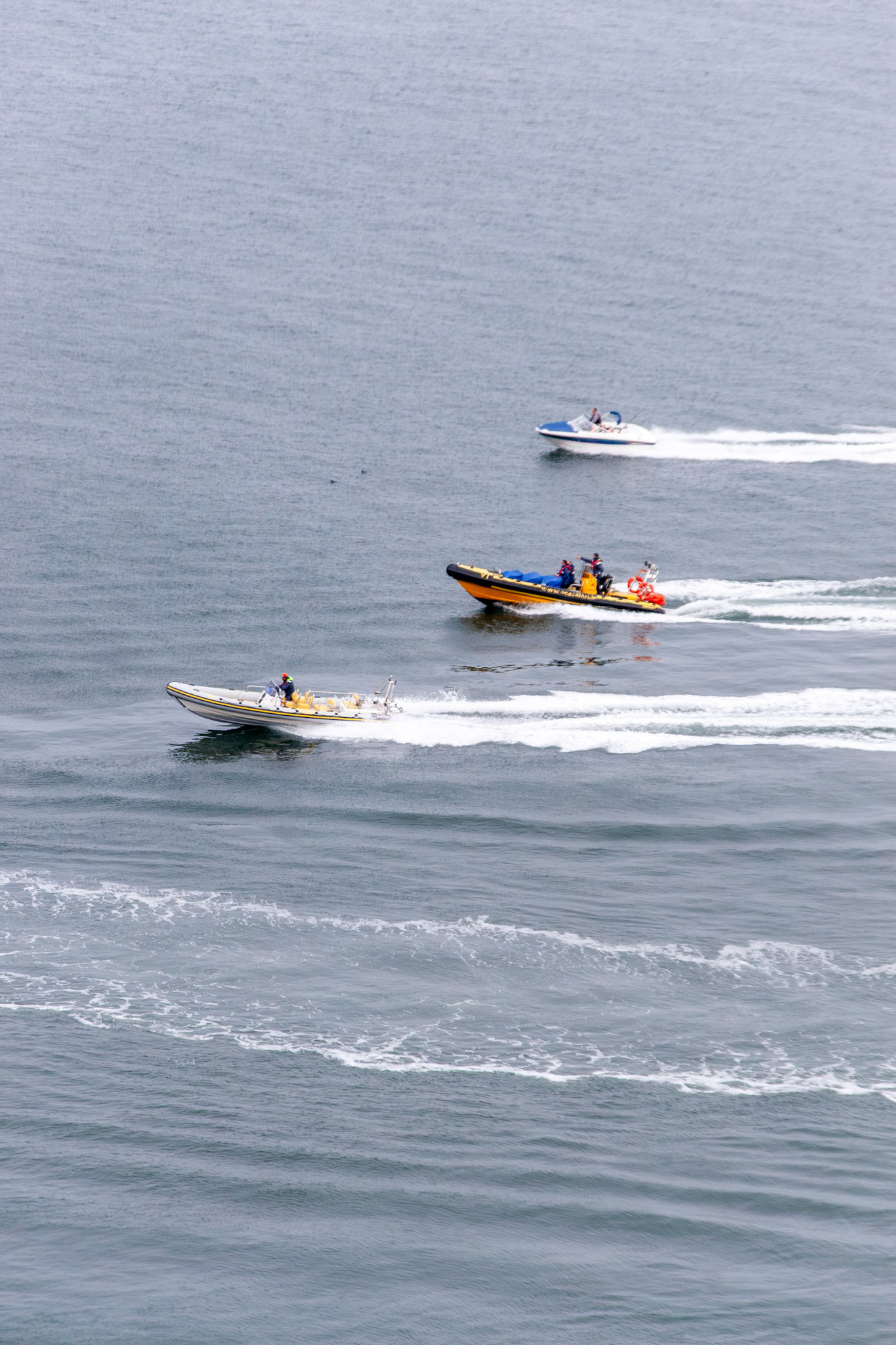 Three high-speed motorboats, including two rigid inflatable boats (RIBs), leave white wakes as they navigate the choppy grey waters of the Irish Sea near Howth, Dublin.