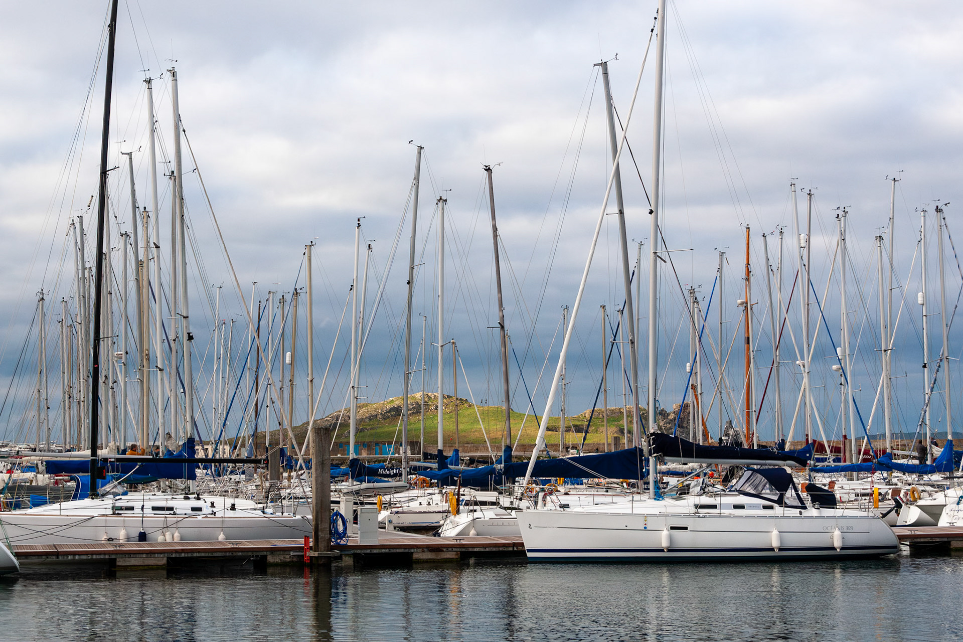 A dense row of modern white sailboats and luxury yachts moored at the docks of Howth Harbor, Dublin, with the scenic green landscape of Ireland's Eye island visible in the background under a cloudy sky.