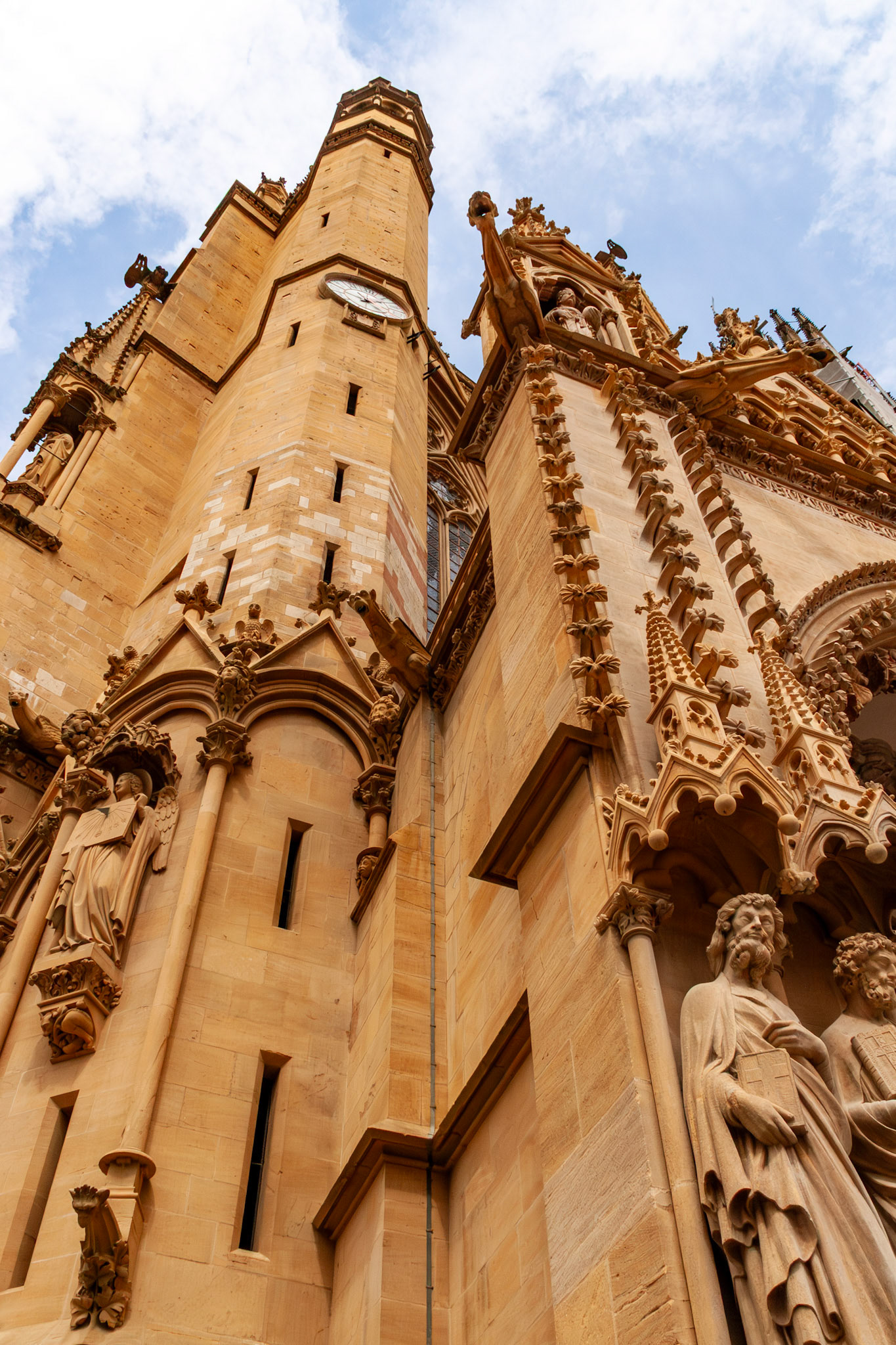 A low-angle architectural shot of the Saint Stephen Cathedral (Cathédrale Saint Étienne) in Metz, France, showcasing the intricate Gothic stonework, religious statues, gargoyles, and the soaring Mutte tower against a blue sky.