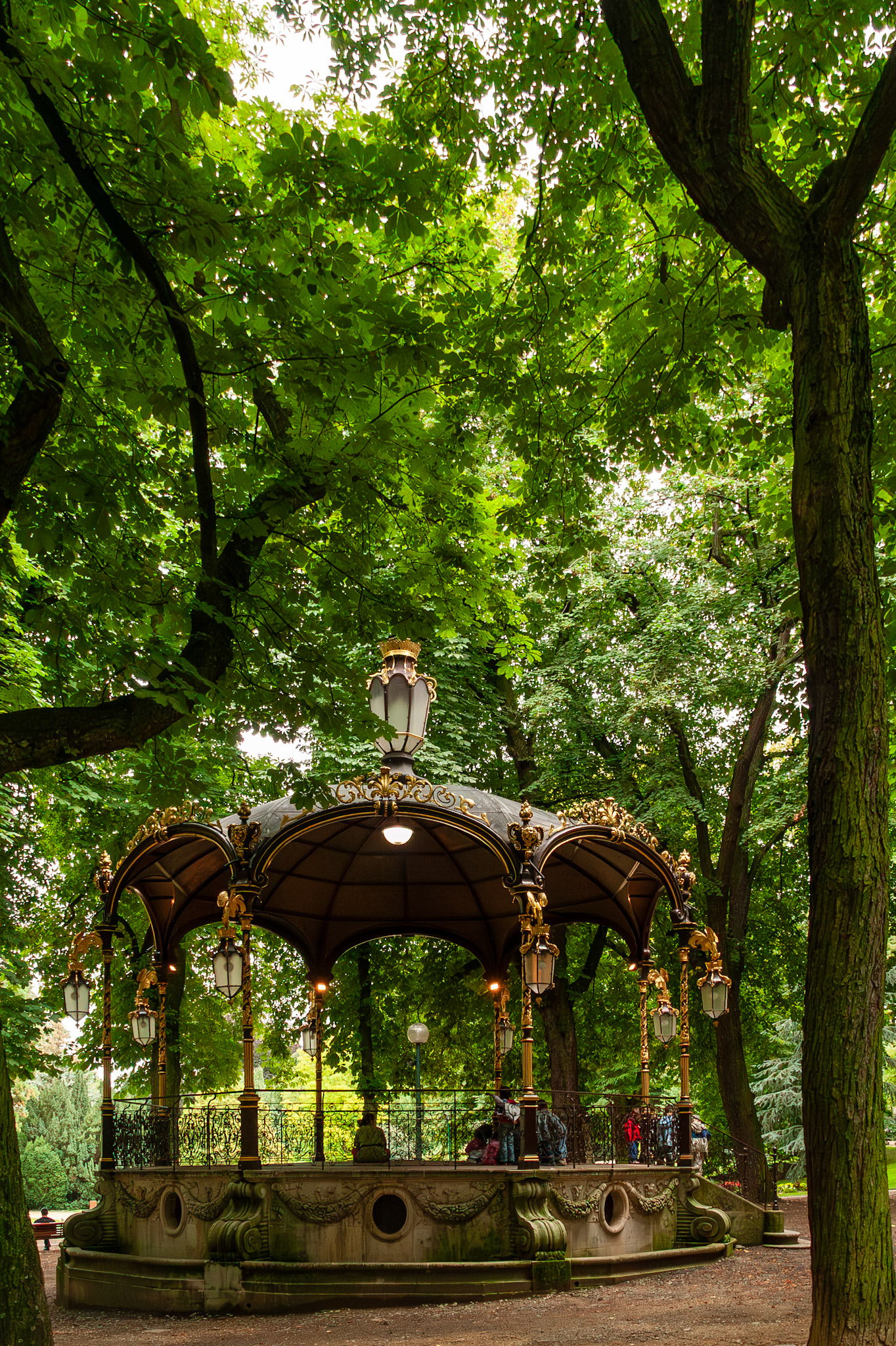 An elegant, 19th-century bandstand or gazebo featuring ornate gold trim and hanging lanterns, nestled among tall, lush green trees within the historic Parc de la Pépinière in Nancy, France.