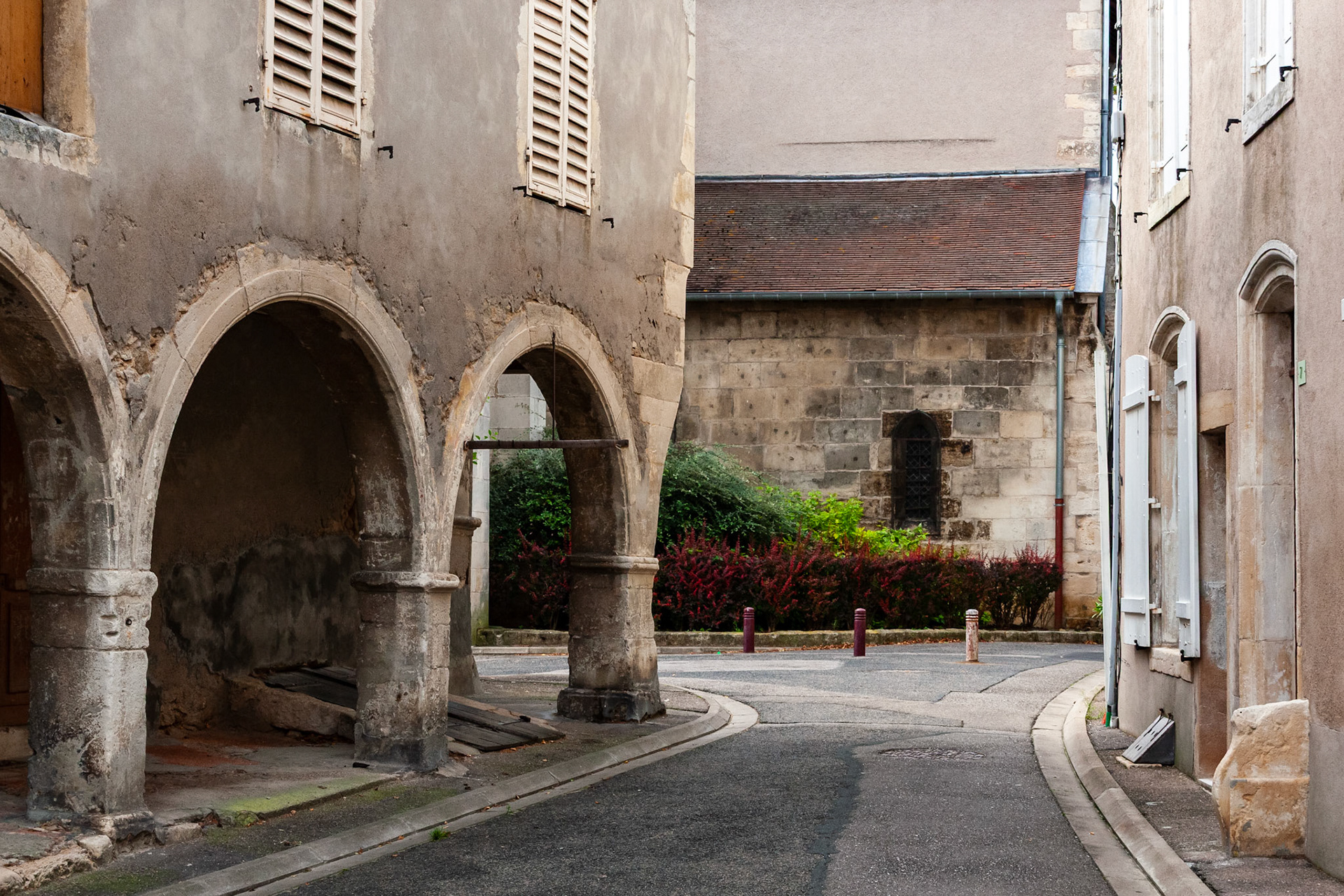 A quiet street scene in a historic French village, featuring weathered stone facades, medieval-style stone arches, and traditional window shutters under soft daylight.