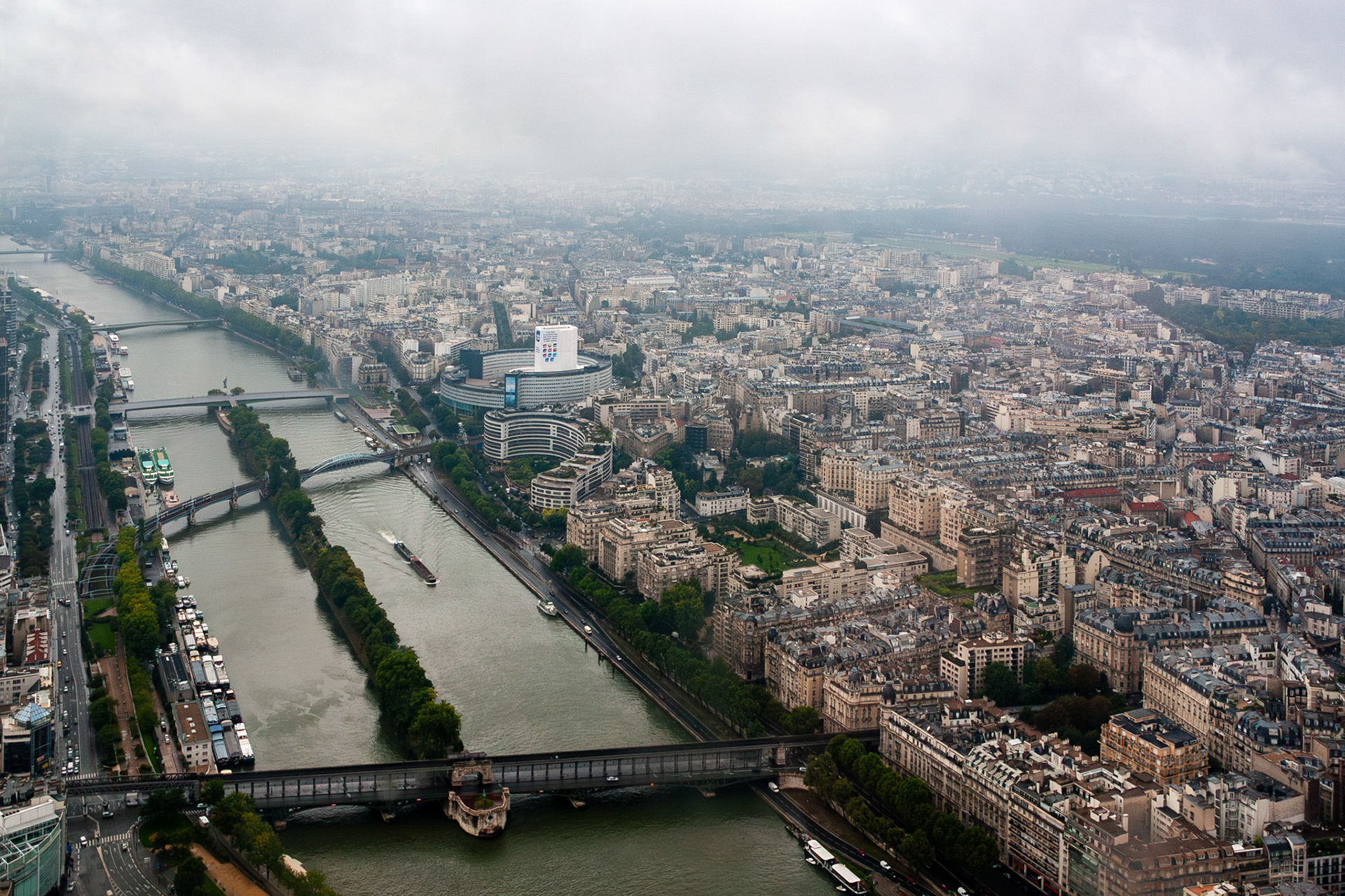 A high-angle panoramic view of the Seine River winding through Paris, France, featuring numerous bridges, houseboats, and the dense urban architecture of the city under a dramatic, overcast sky.