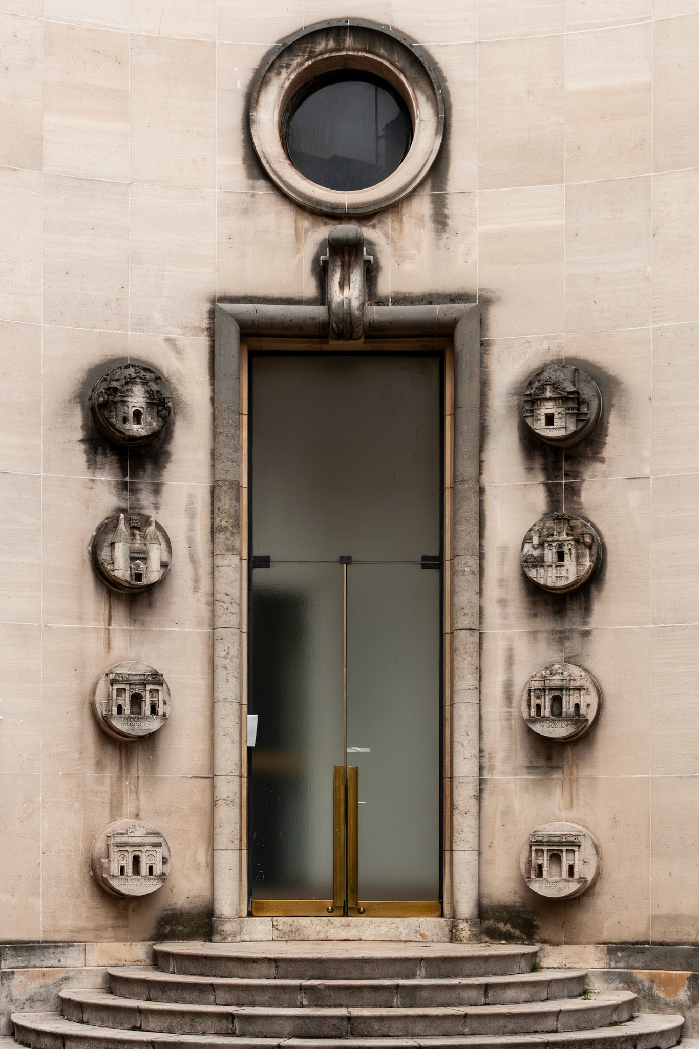 A symmetrical view of a stone building entrance in Nancy, France, featuring a tall frosted glass door framed by eight circular stone medallions depicting various architectural landmarks and a round oculus window above.