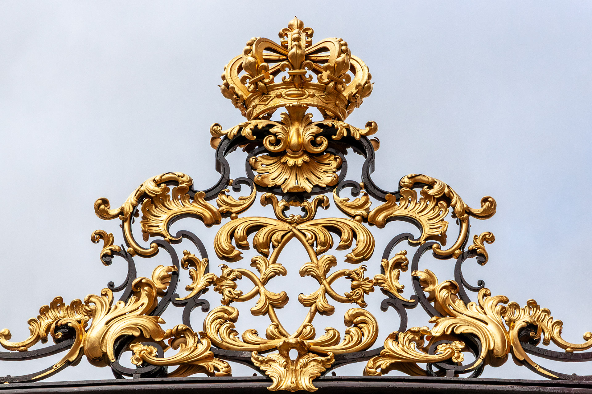 A detailed close-up of a magnificent gold-leafed crown ornament atop intricate black and gold wrought iron gates, showcasing the grand Rococo architectural style of Place Stanislas in Nancy, France.