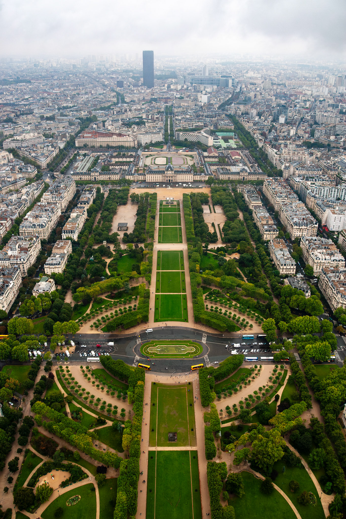 A stunning vertical high-angle shot looking down the symmetrical axis of the Champ de Mars toward the École Militaire and the Montparnasse Tower, showcasing Parisian urban planning under a cloudy sky.