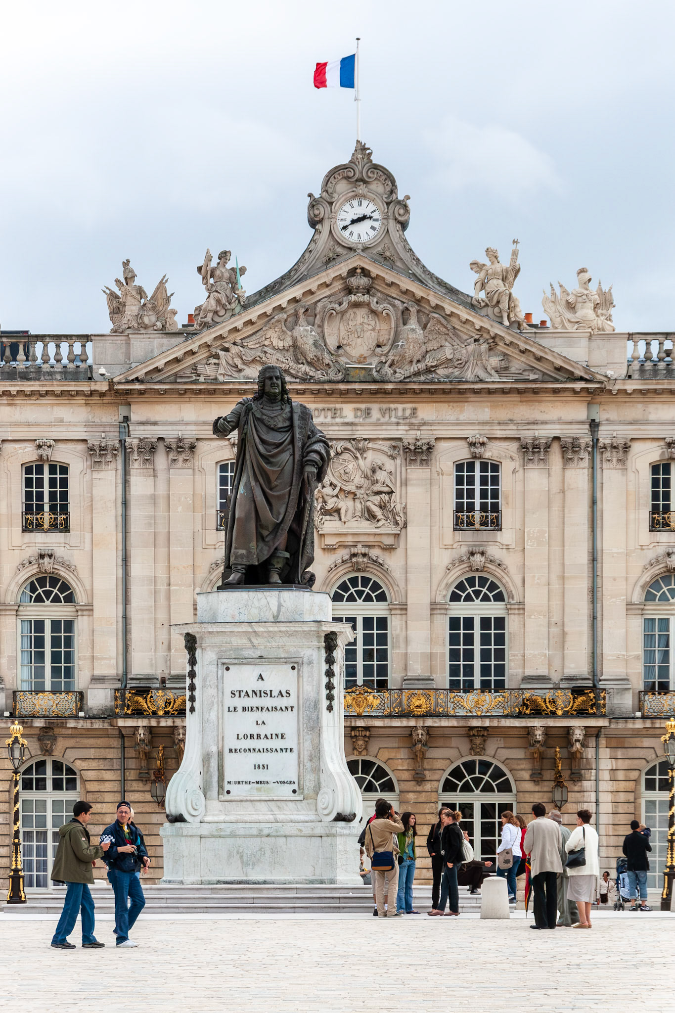 A full view of the bronze statue of Stanislas Leszczynski in the center of Place Stanislas, with the grand neoclassical facade of the Nancy City Hall (Hôtel de Ville) and a waving French flag in the background.