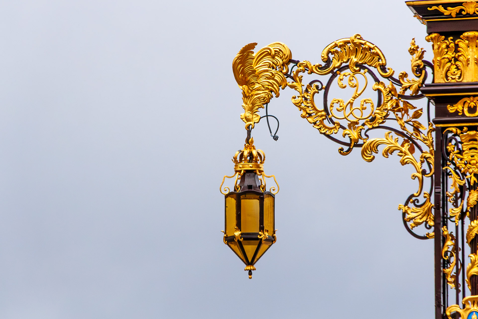 A detailed close-up of an exquisite, gold-leafed wrought iron street lamp featuring intricate scrollwork and a rooster emblem, set against a neutral sky in Nancy, France.