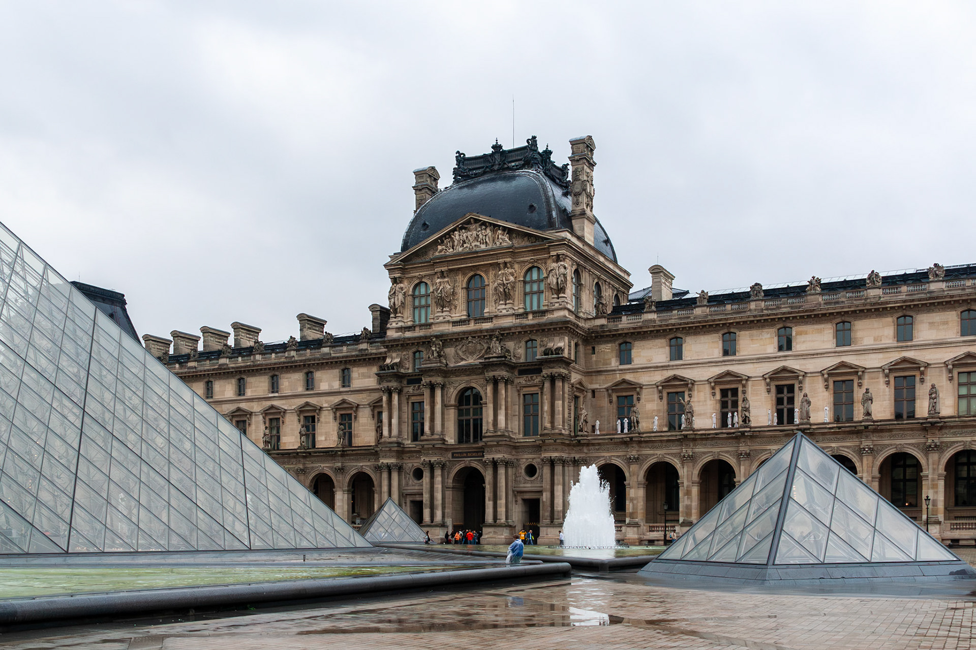 A striking juxtaposition of modern glass pyramid structures and a grand Renaissance-style palace facade in a Parisian courtyard. The scene includes water fountains and classical stone sculptures under an overcast sky.