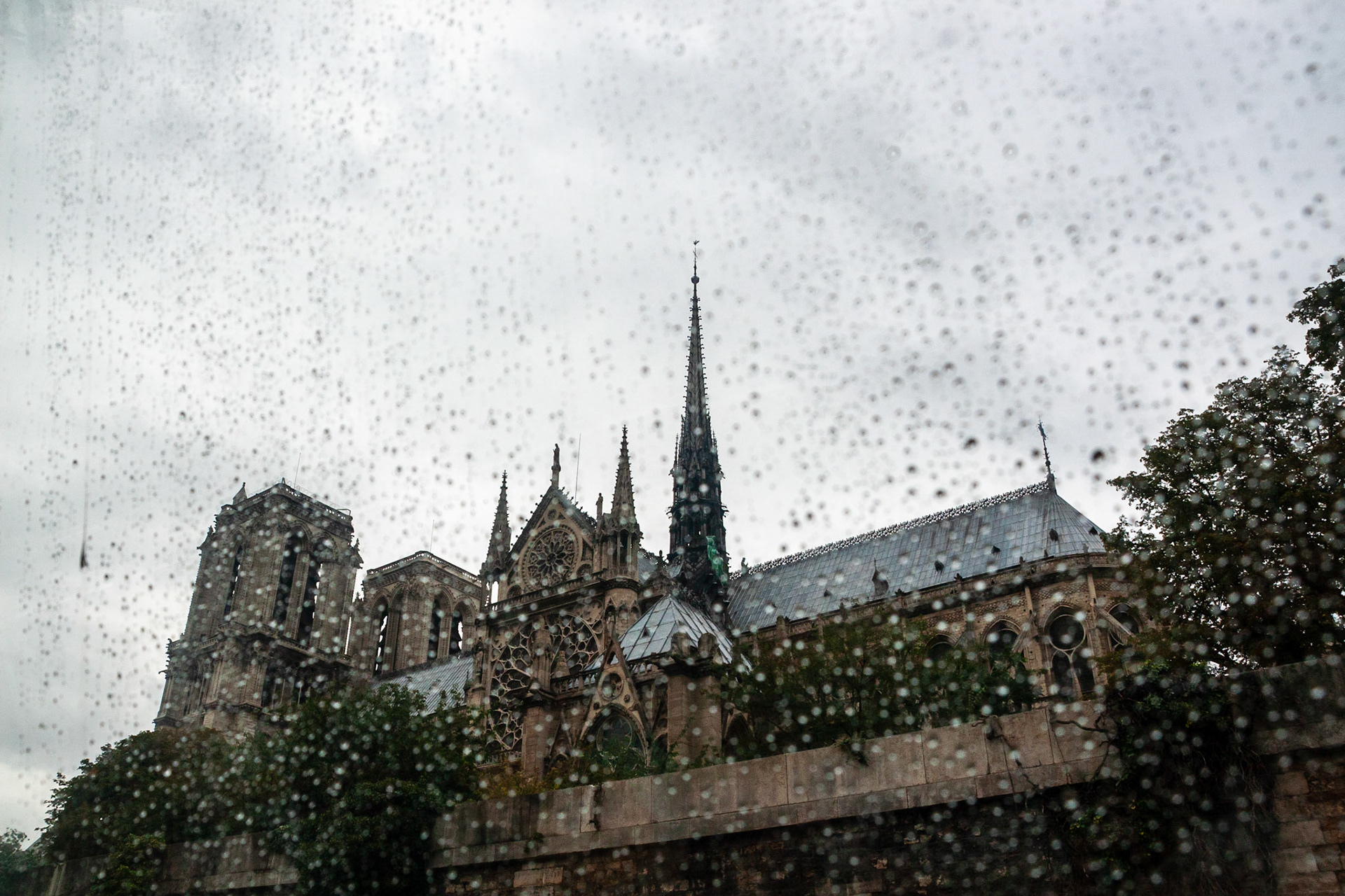 The Gothic architecture of the Notre Dame de Paris cathedral, featuring its iconic spire and rose window, viewed through a rain-covered window pane that creates a textured, moody urban atmosphere.