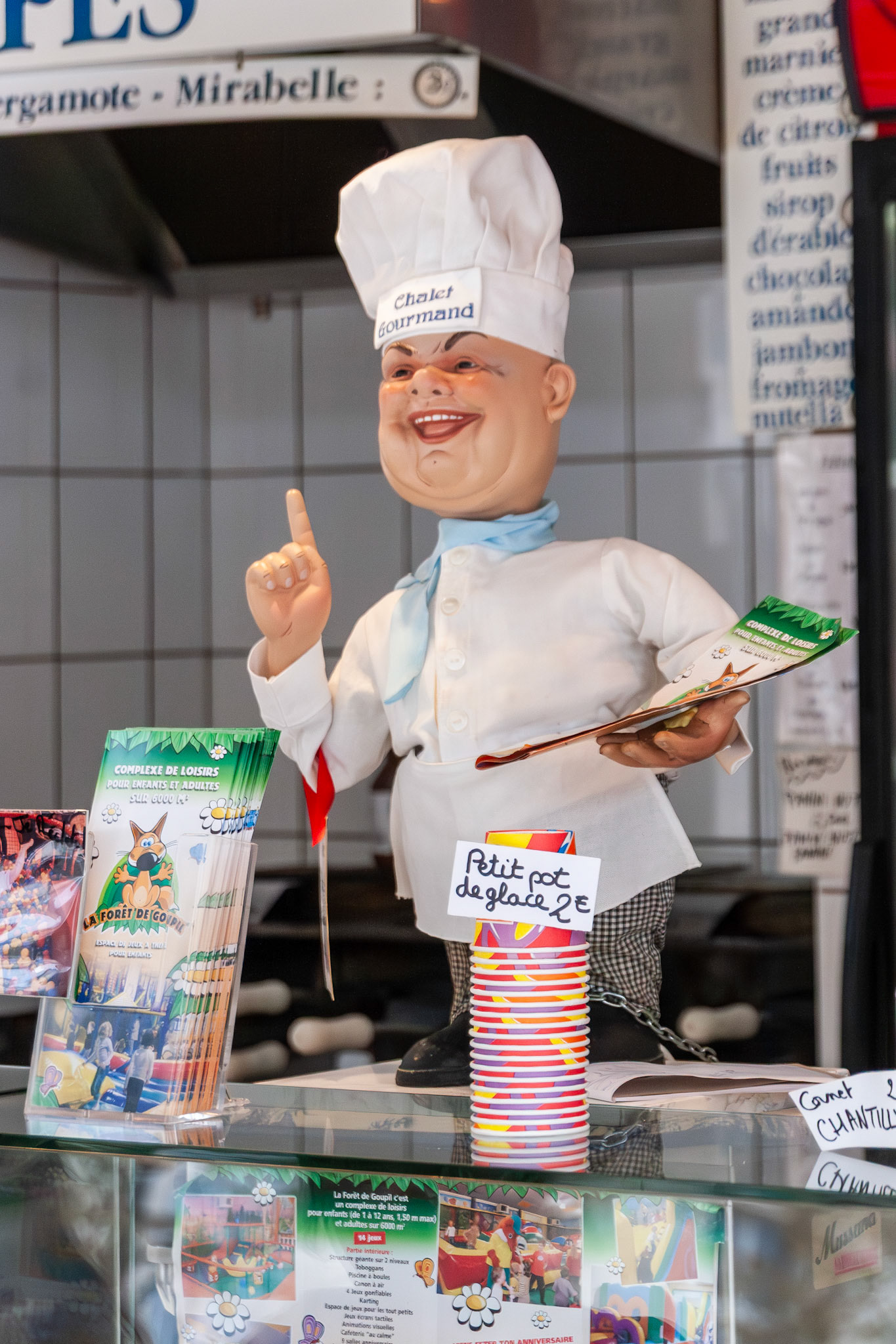 A playful "Chalet Gourmand" chef figurine stands on a glass counter at an outdoor food stall, surrounded by colorful menus and stacked ice cream cups in Nancy, France.