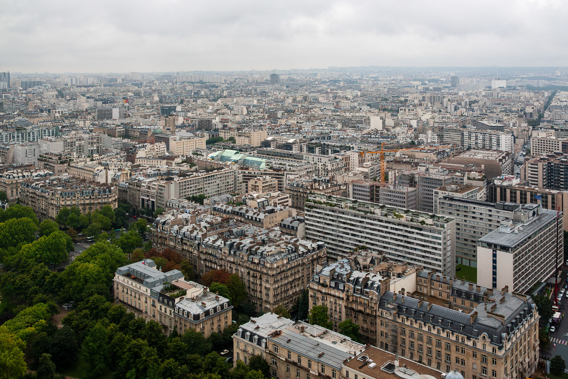 An expansive aerial perspective of the dense urban fabric of Paris, France, showcasing traditional Haussmann-style buildings, green parks, and modern city infrastructure under an overcast sky.