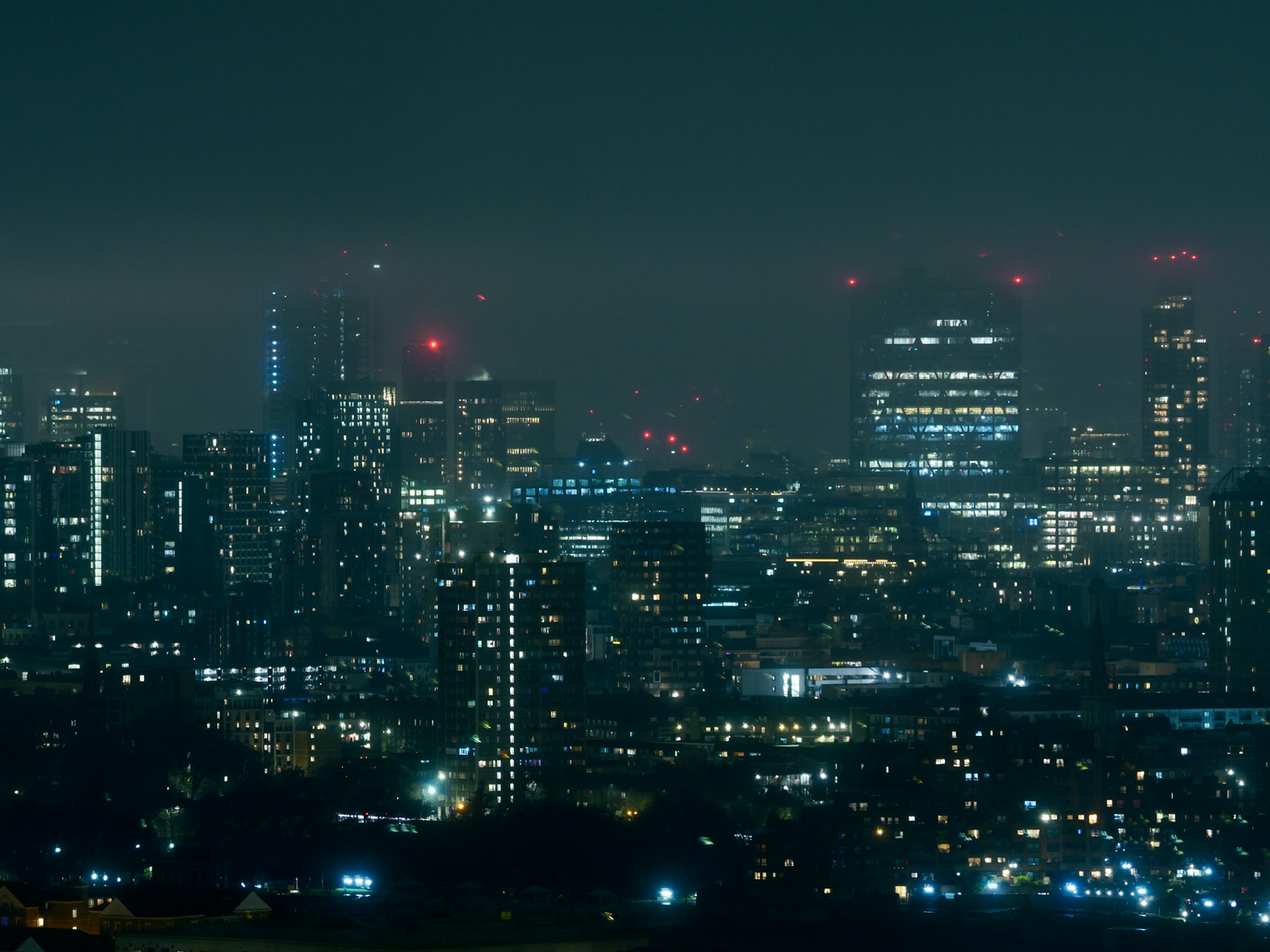 A high-angle, atmospheric night view of the London cityscape featuring illuminated skyscrapers, construction cranes, and glowing red aviation lights under a misty, dark sky, capturing a moody cyberpunk-inspired urban environment.